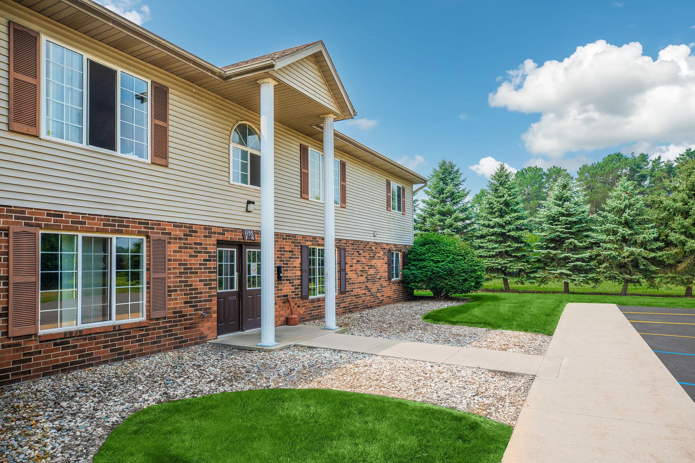 A two-story brick building with large windows and white columns at the entrance, surrounded by well-maintained grass and a landscaped area. In the background, there are tall green trees and a clear blue sky with a few clouds. The path leading to the entrance is made of concrete.