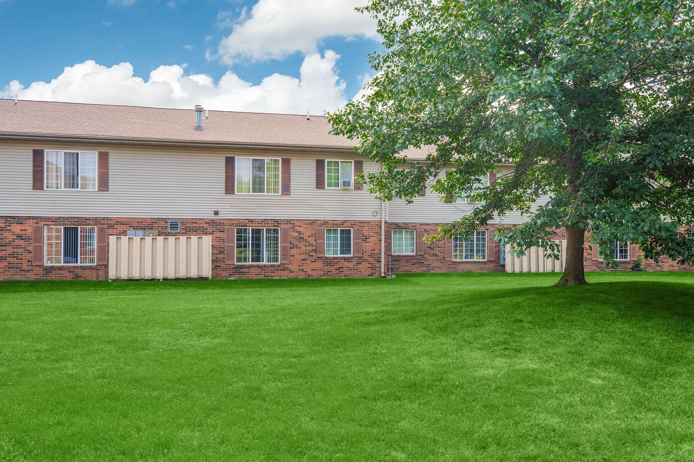 A view of a two-story residential building with multiple windows, set against a bright blue sky with white clouds. A large, lush green lawn is in the foreground, and a tree is situated nearby, adding to the outdoor aesthetic of the property.