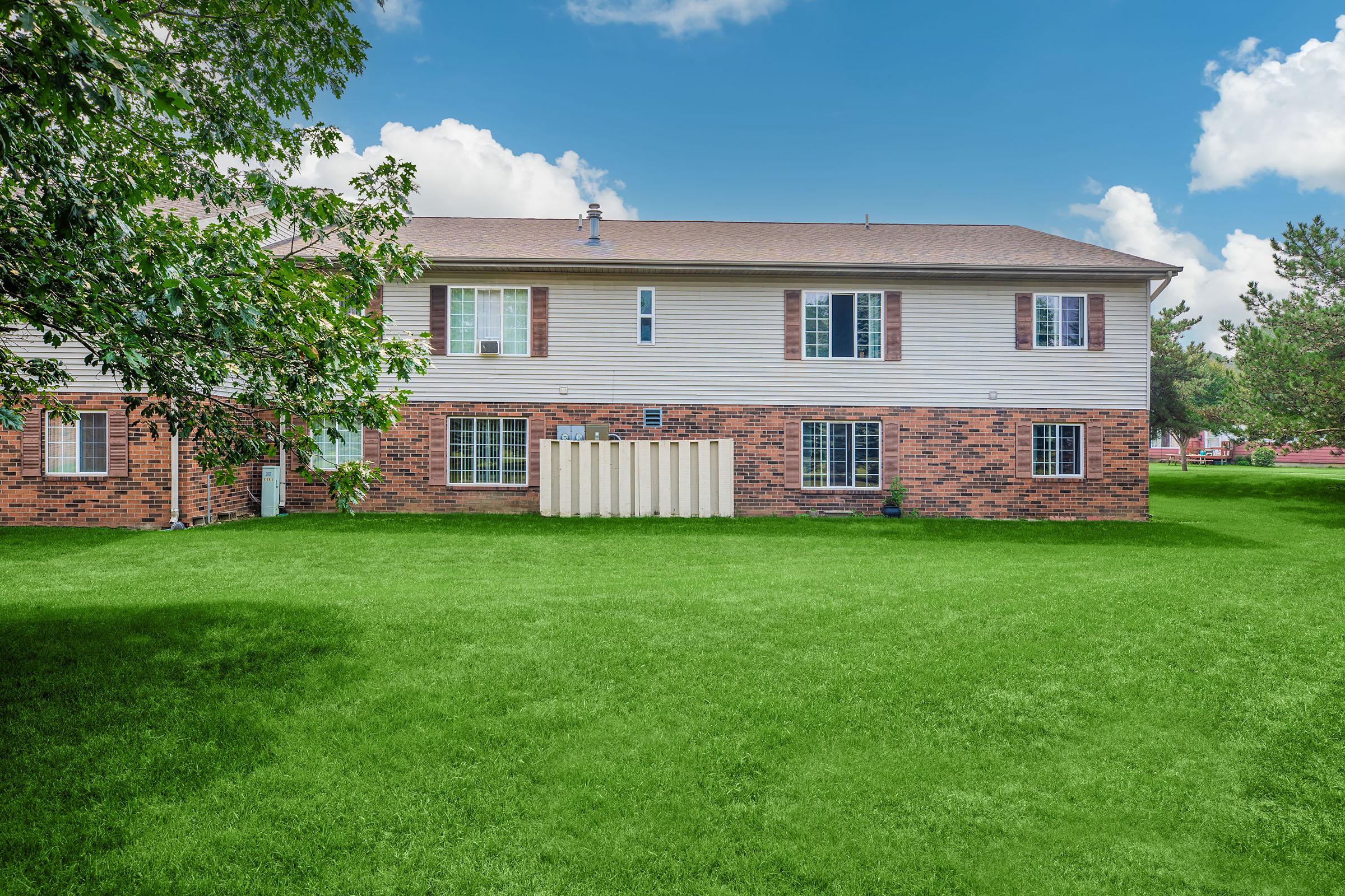 A well-maintained brick and siding apartment building with multiple windows, situated on a lush green lawn. The sky is clear with a few clouds, creating a bright and inviting atmosphere. There is a small storage area visible in the yard.