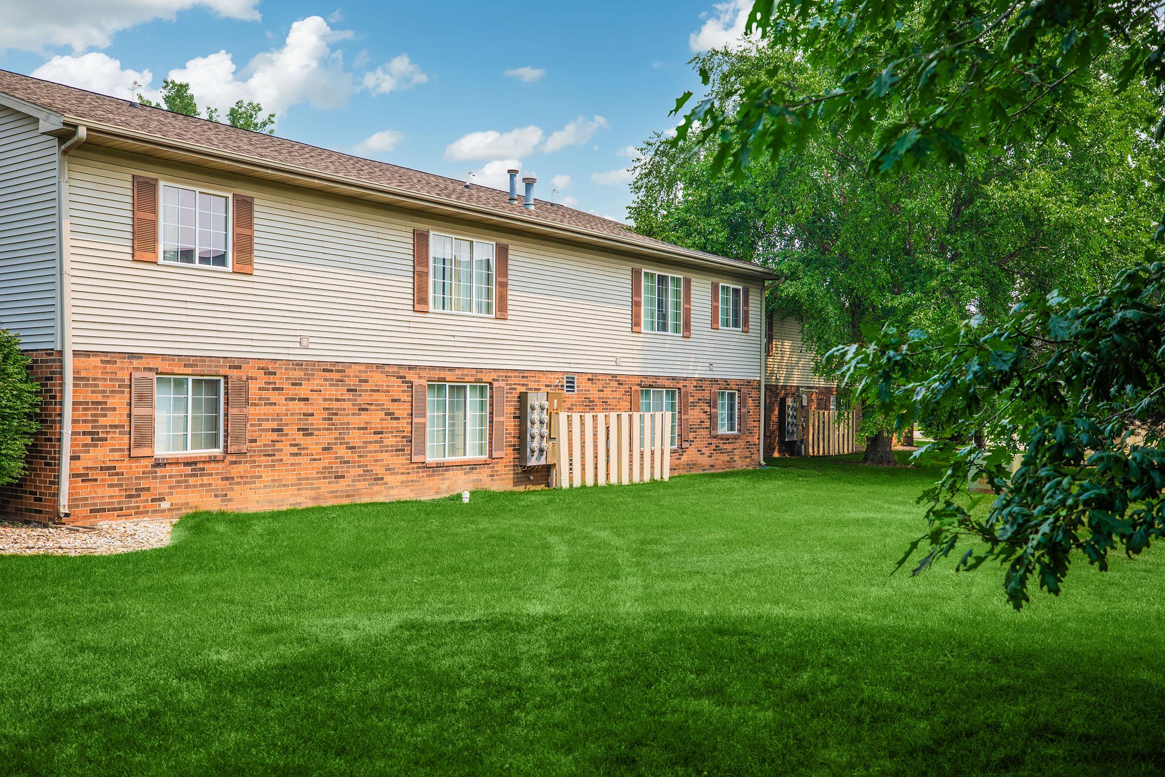 A two-story residential building with a brick and siding exterior, featuring several windows. The property includes a neatly maintained green lawn, trees, and a clear blue sky with fluffy clouds in the background. The scene conveys a tranquil neighborhood atmosphere.