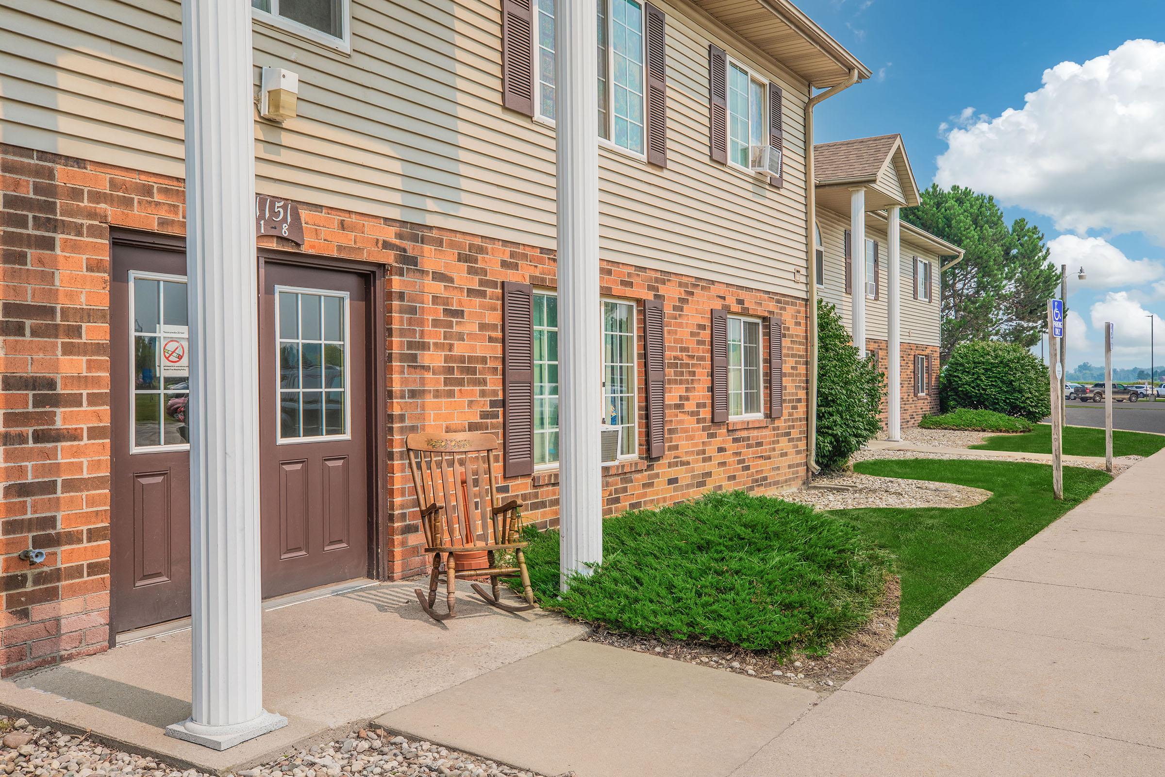 Exterior view of a residential building with brick and siding, featuring a front porch with a wooden chair. Green shrubs line the walkway, and the area is under a clear blue sky with a few clouds.