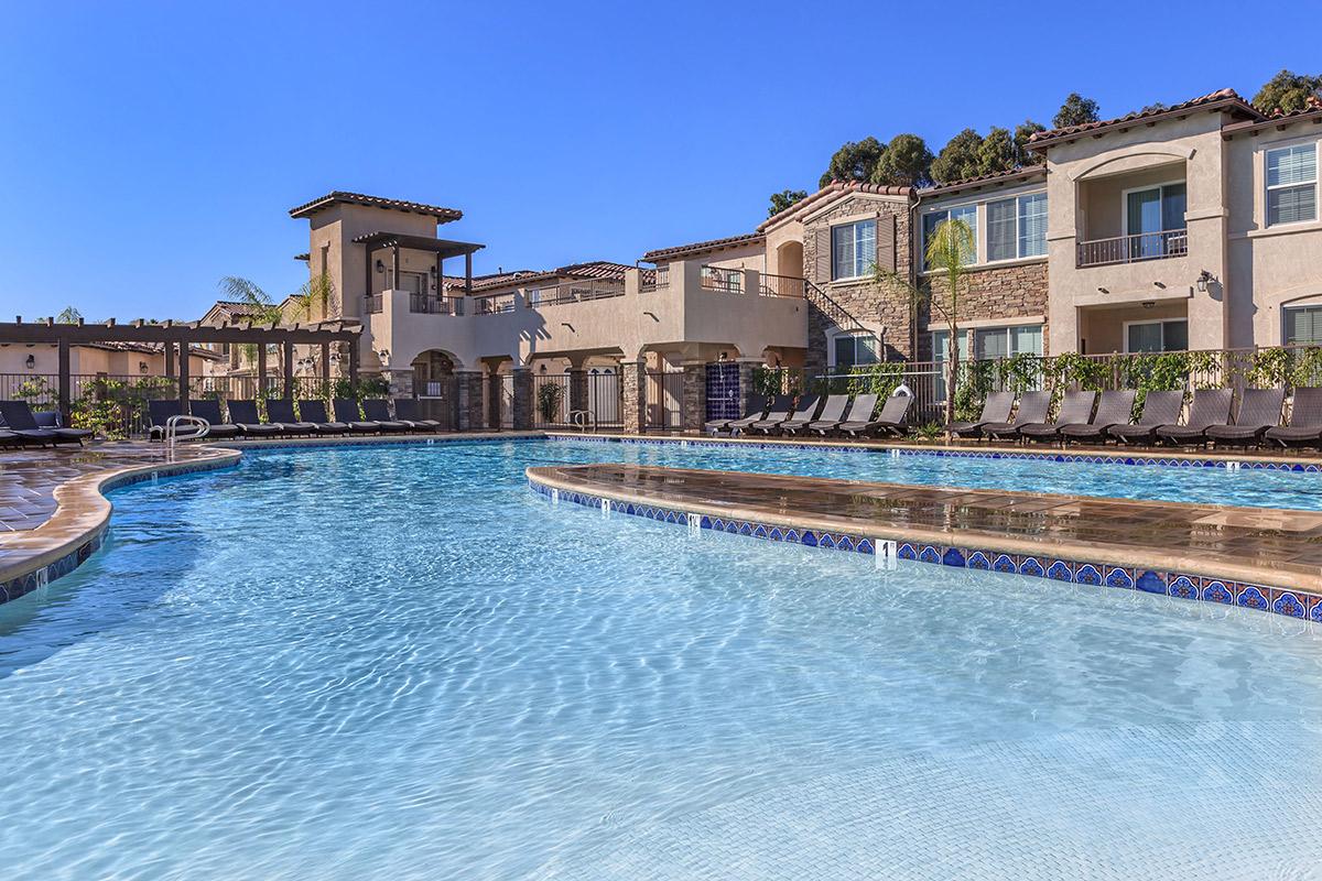 A clear blue swimming pool surrounded by lounge chairs, with a stylish pool deck and palm trees in the background. Modern buildings line the pool area, showcasing a sunny day with a blue sky, creating a resort-like atmosphere.