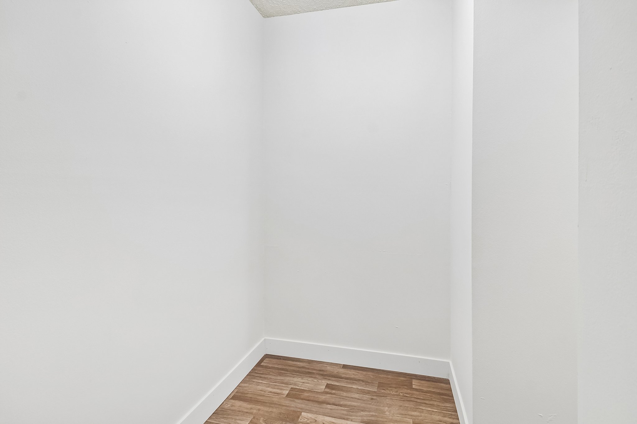 A small, empty room with white walls and a wooden floor. The space has no furniture or decorations and features a ceiling that is out of view. It appears to be a closet or storage area, providing a minimalistic and clean appearance.