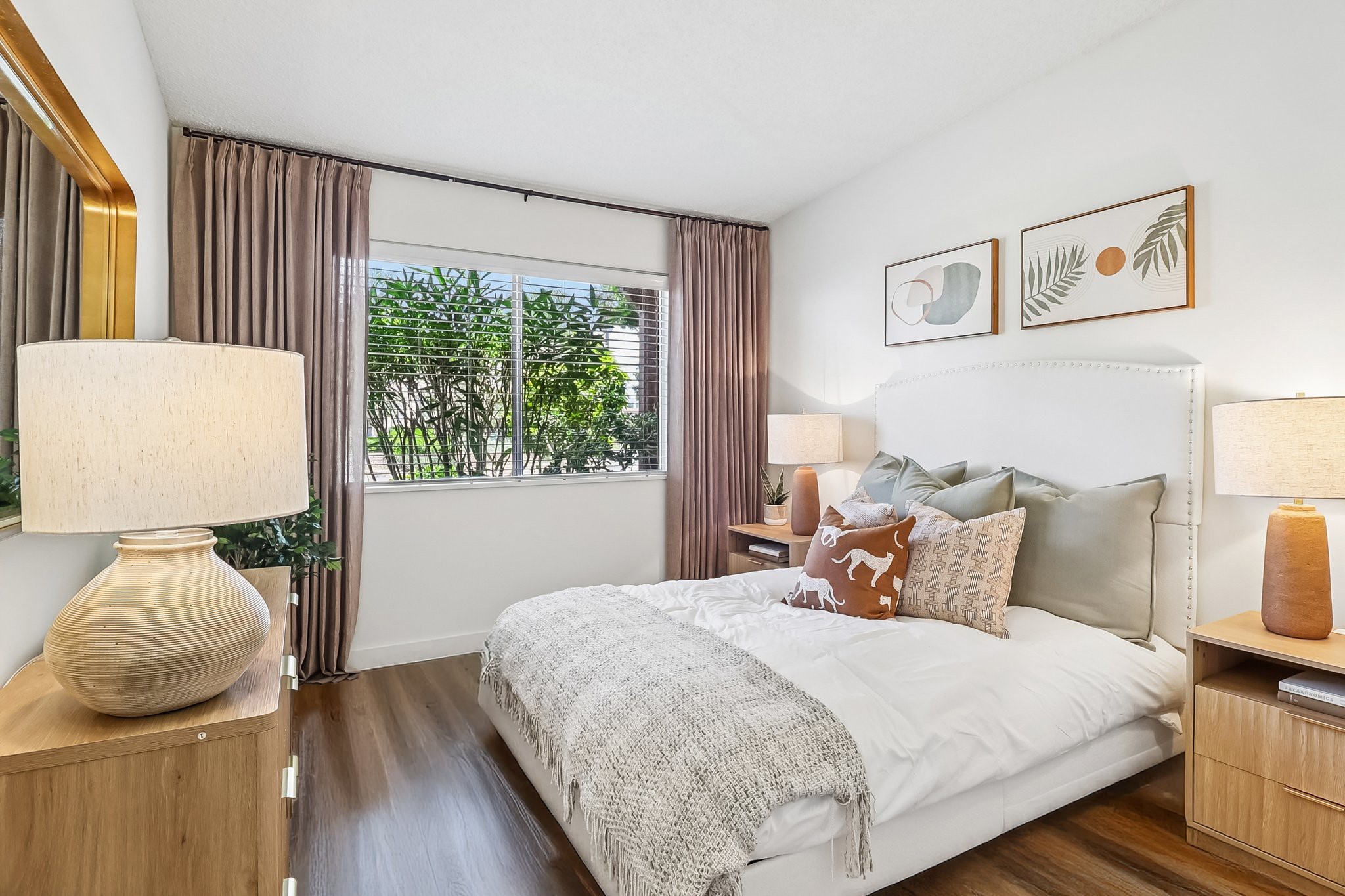 Cozy bedroom featuring a large window with soft curtains, a comfortable white bed adorned with decorative pillows and a throw blanket, two bedside lamps, and a wooden dresser. The decor includes framed artwork and greenery visible outside the window, creating a warm and inviting atmosphere.