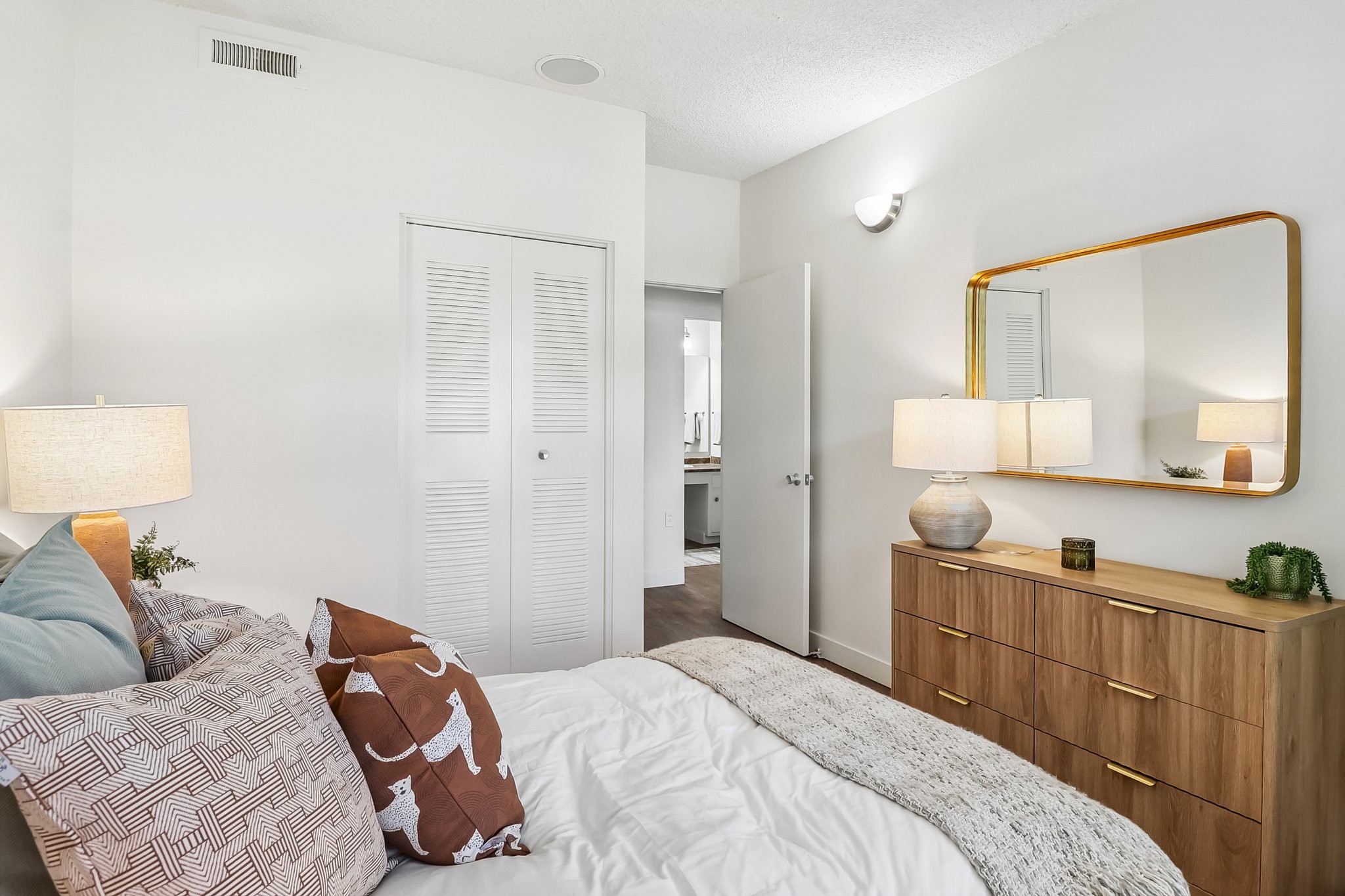 Cozy bedroom featuring a bed with decorative pillows, a wooden dresser, and a large mirror. Soft lighting from bedside lamps and a bright, minimalist decor enhance the inviting atmosphere. A door leads to a bathroom, and the room has light-colored walls with accents of greenery.