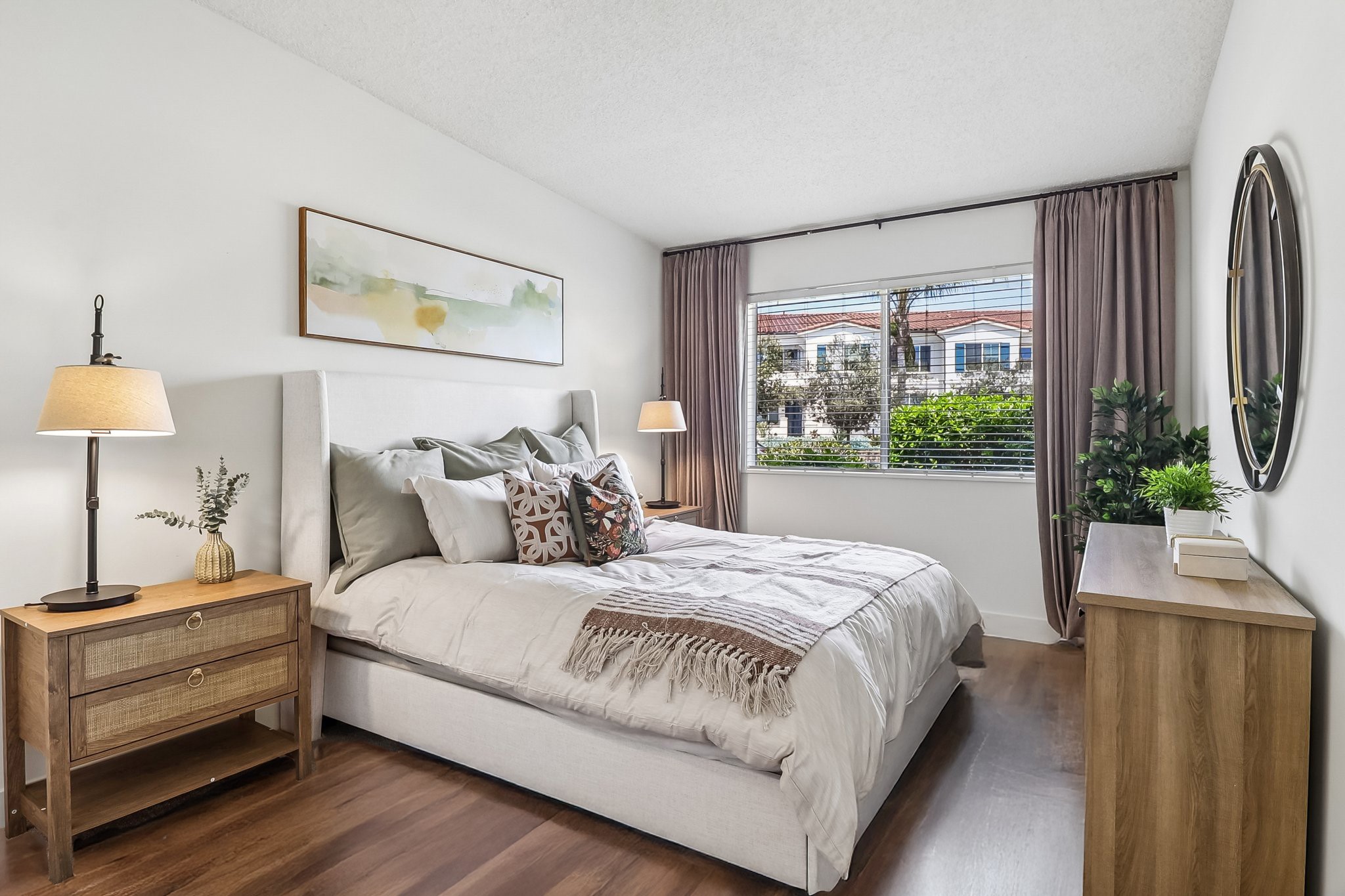 A cozy bedroom featuring a light-colored bed with decorative pillows, a warm-toned blanket, and two bedside tables with lamps. A large window fills the room with natural light, showcasing greenery outside. Soft curtains frame the window, and a wall mirror adds depth to the space.