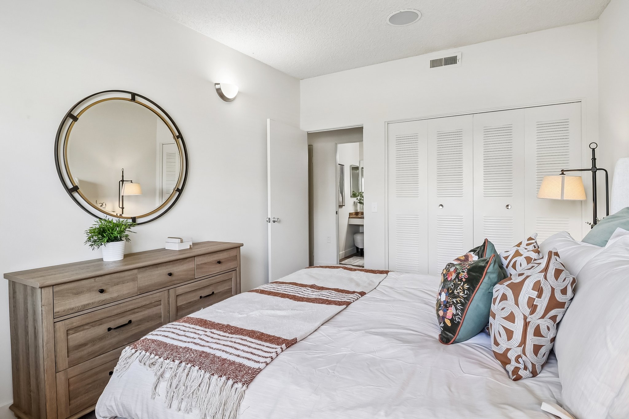 A cozy bedroom featuring a large round mirror above a wooden dresser. The bed is adorned with decorative pillows and a patterned throw blanket. White walls and closets create a minimalist aesthetic, while a lamp beside the bed adds a warm touch. Natural light illuminates the space, enhancing its inviting atmosphere.