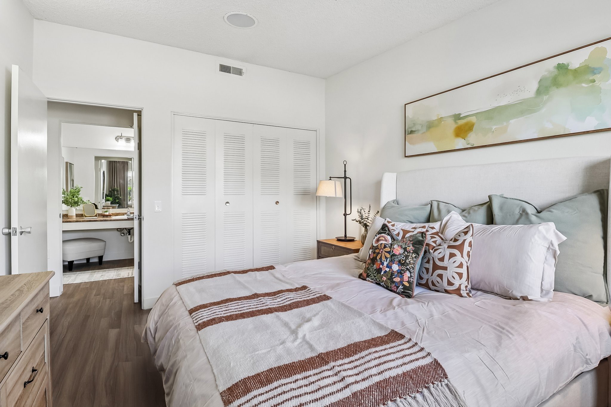 Cozy bedroom featuring a large bed with decorative pillows, light-colored bedding, and a bedside lamp. A piece of abstract art hangs above the bed. The room includes a closet with louvered doors and an open doorway leading to a bathroom with a modern vanity and mirror, complemented by plants for a fresh touch.