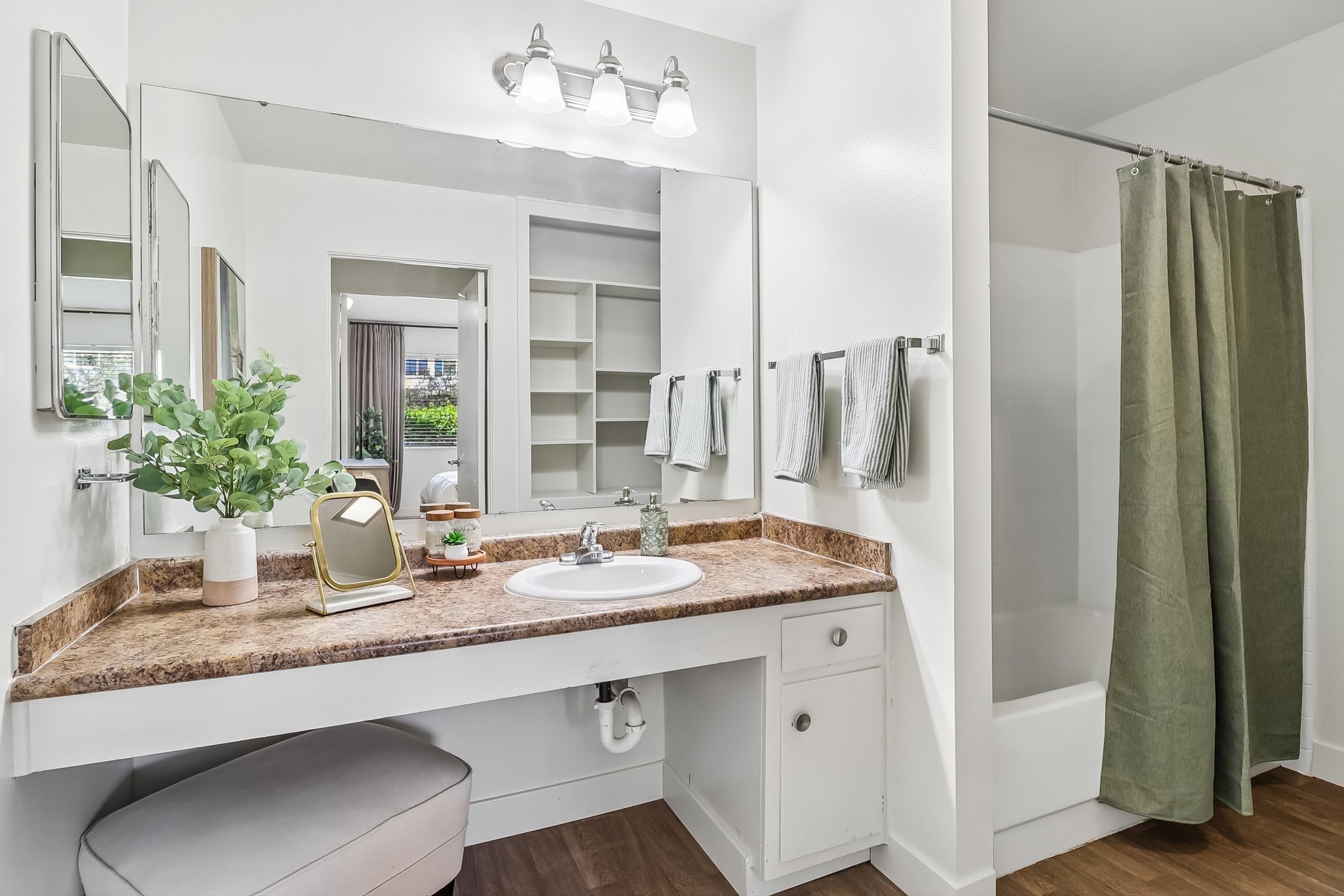 A modern bathroom featuring a double sink vanity with a large mirror and decorative plants. The countertop is adorned with toiletries, and there are neatly arranged towels hanging nearby. A shower curtain in soft green separates the shower area, and light-colored walls create a bright, airy atmosphere.