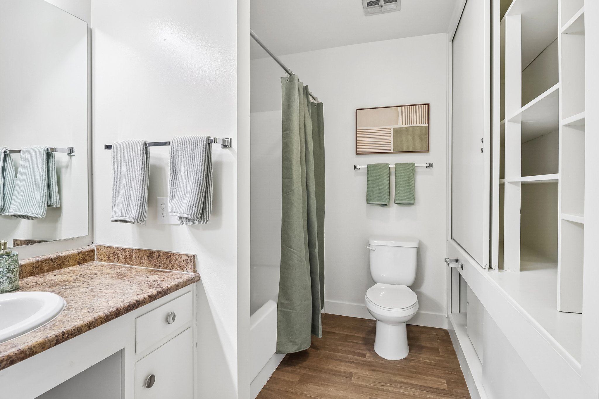 A modern bathroom featuring a white toilet, a shower with a green curtain, a sink with a granite countertop, and a wall-mounted mirror. Light-colored walls and wooden flooring create a spacious feel, complemented by green towels and decor. Shelving is visible next to the toilet.