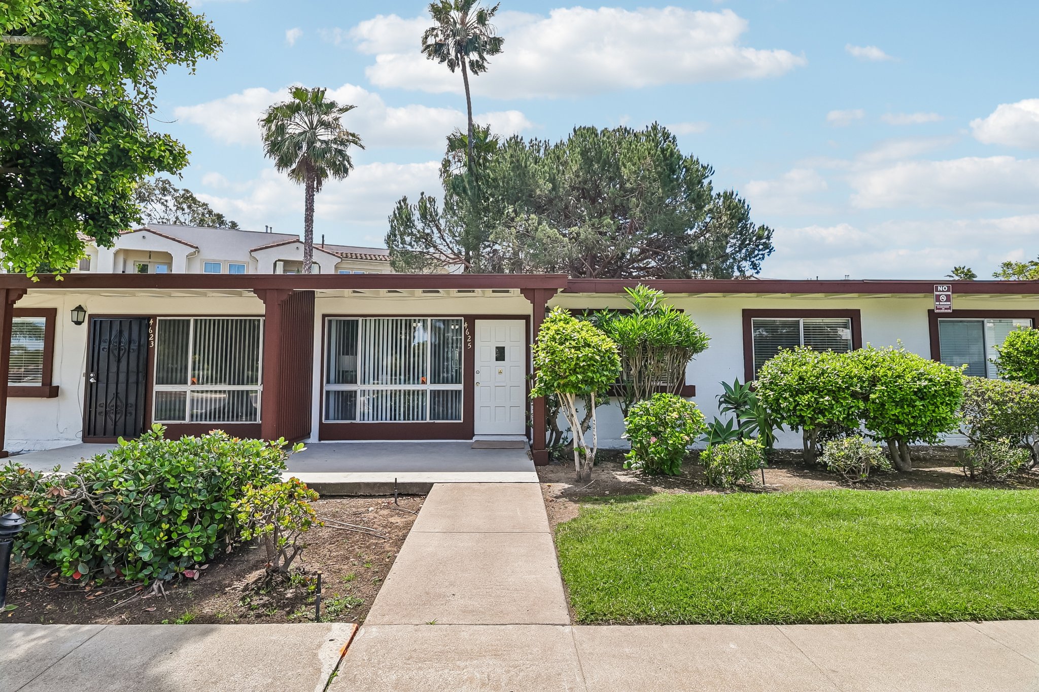 A charming single-story residential building with a white facade and red trim. The entrance features a white door framed by large windows. Lush green bushes line the pathway, and palm trees are visible in the background under a partly cloudy sky, creating a welcoming outdoor space.