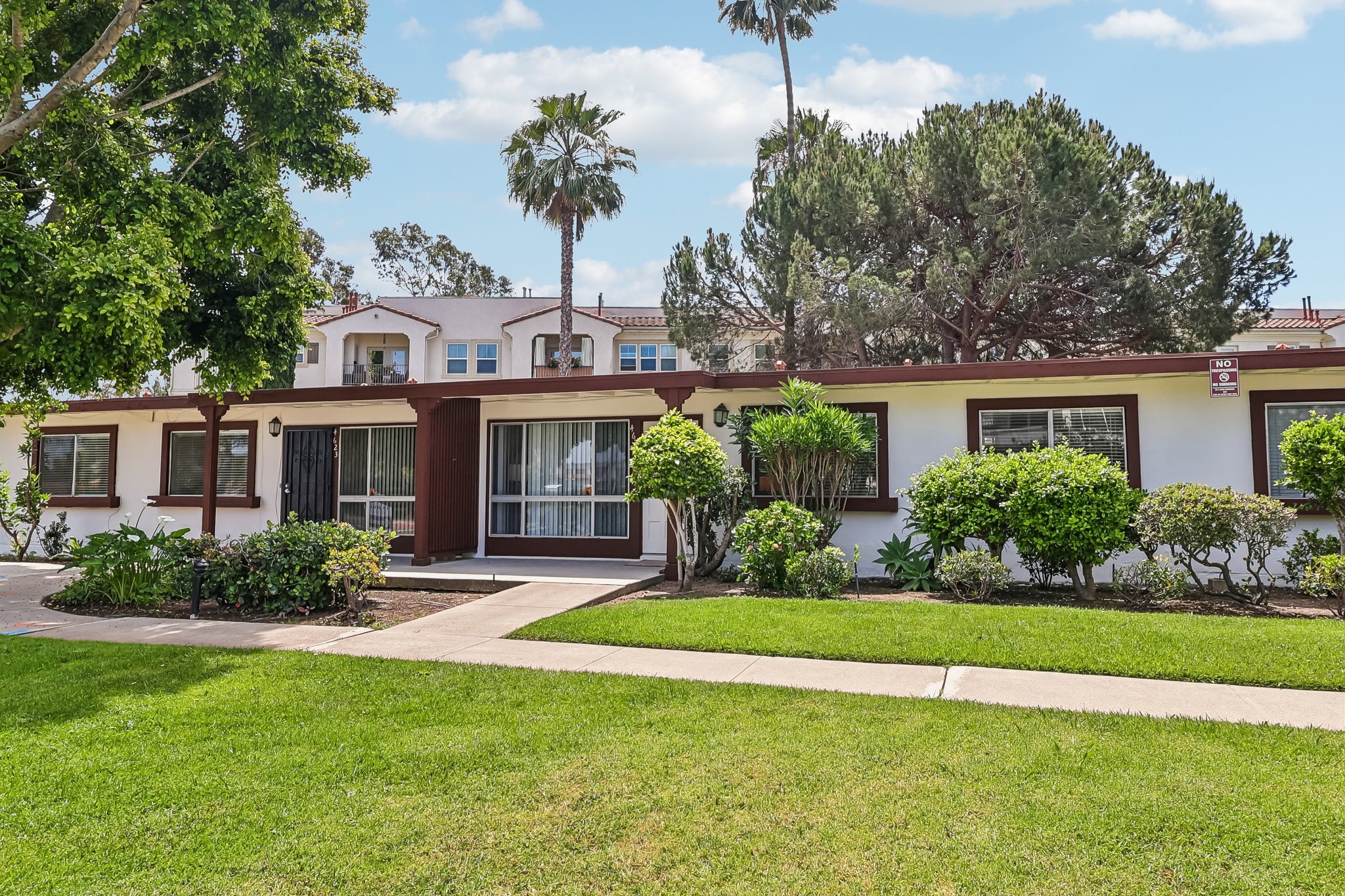A well-maintained apartment building surrounded by lush green grass and landscaping. The building features a facade with white walls, brown trim, and several palm trees in the background, creating a welcoming atmosphere. Bright blue skies with scattered clouds are visible above.