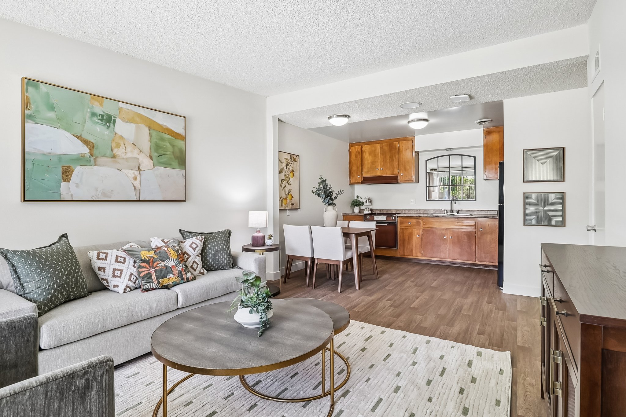 A stylish living area featuring a light gray sofa with decorative pillows, a round coffee table, and a modern artwork on the wall. The open layout includes a dining area with white chairs and a kitchen with wooden cabinets in the background, illuminated by overhead lights.