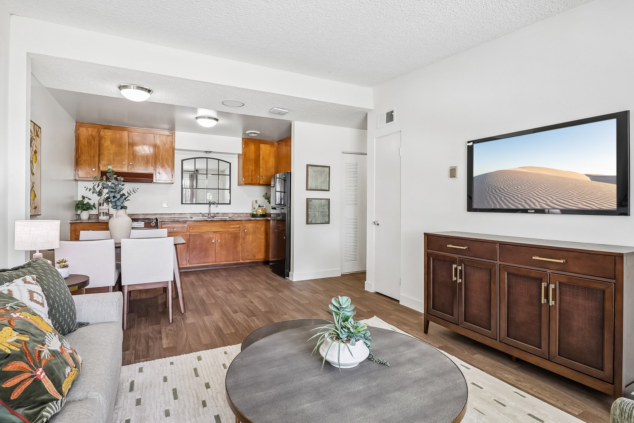 A bright, modern living space featuring a cozy gray sofa with decorative cushions, a round coffee table with a plant, a wooden dining table with chairs, and a kitchen area with wooden cabinetry. A wall-mounted TV displays a desert landscape image. The decor is sleek and inviting.