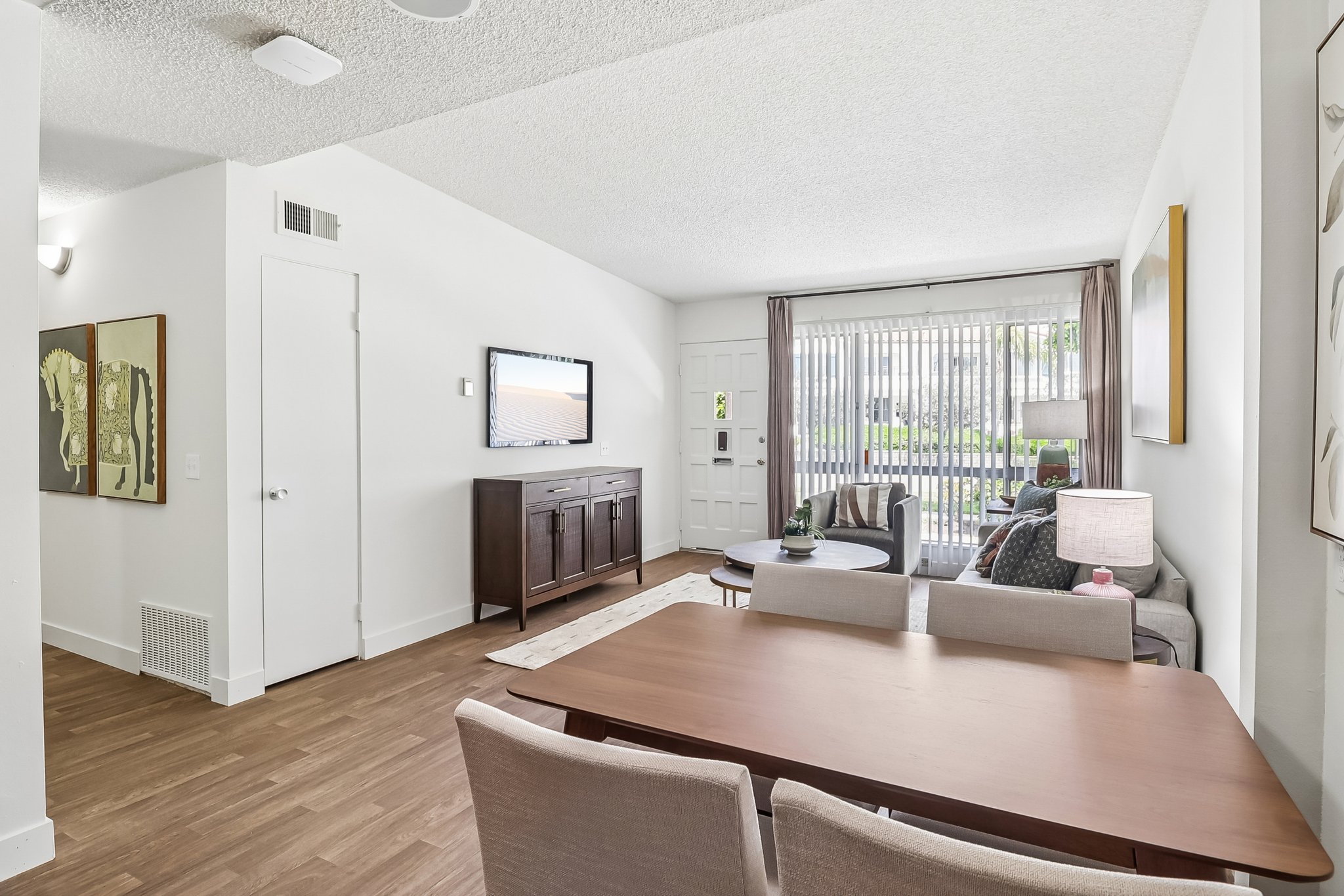 Modern living room and dining area featuring a wooden table with chairs, a comfortable sofa, a coffee table, and a media console with a TV. Large windows with blinds allow natural light, and artwork adorns the walls, creating a cozy and welcoming atmosphere.
