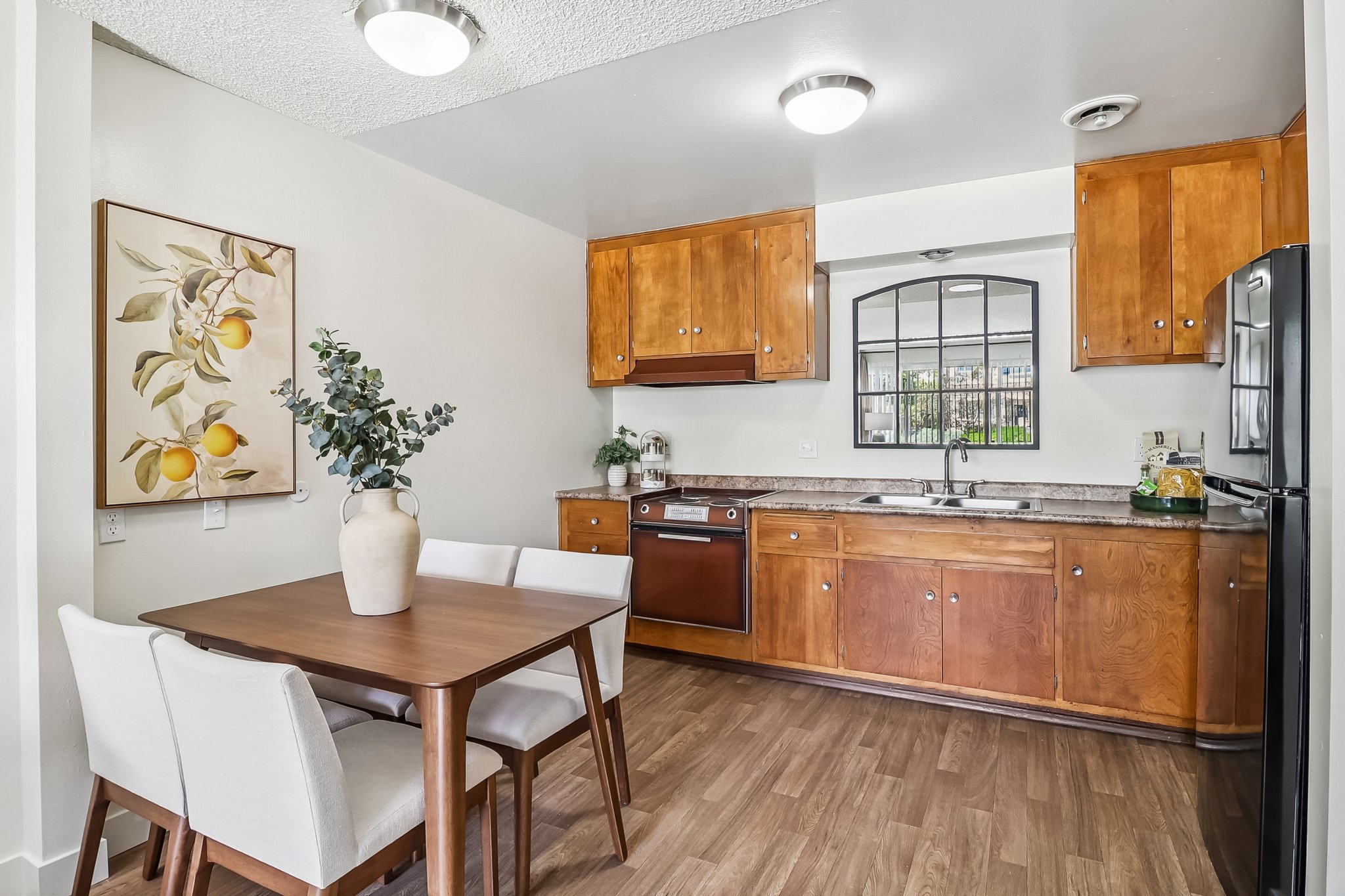 A modern kitchen featuring wooden cabinets, a stainless steel dishwasher, and a black refrigerator. A dining table with four white chairs is set nearby, complemented by a decorative vase with greenery. A framed artwork of lemons hangs on the wall, adding a touch of color to the space.