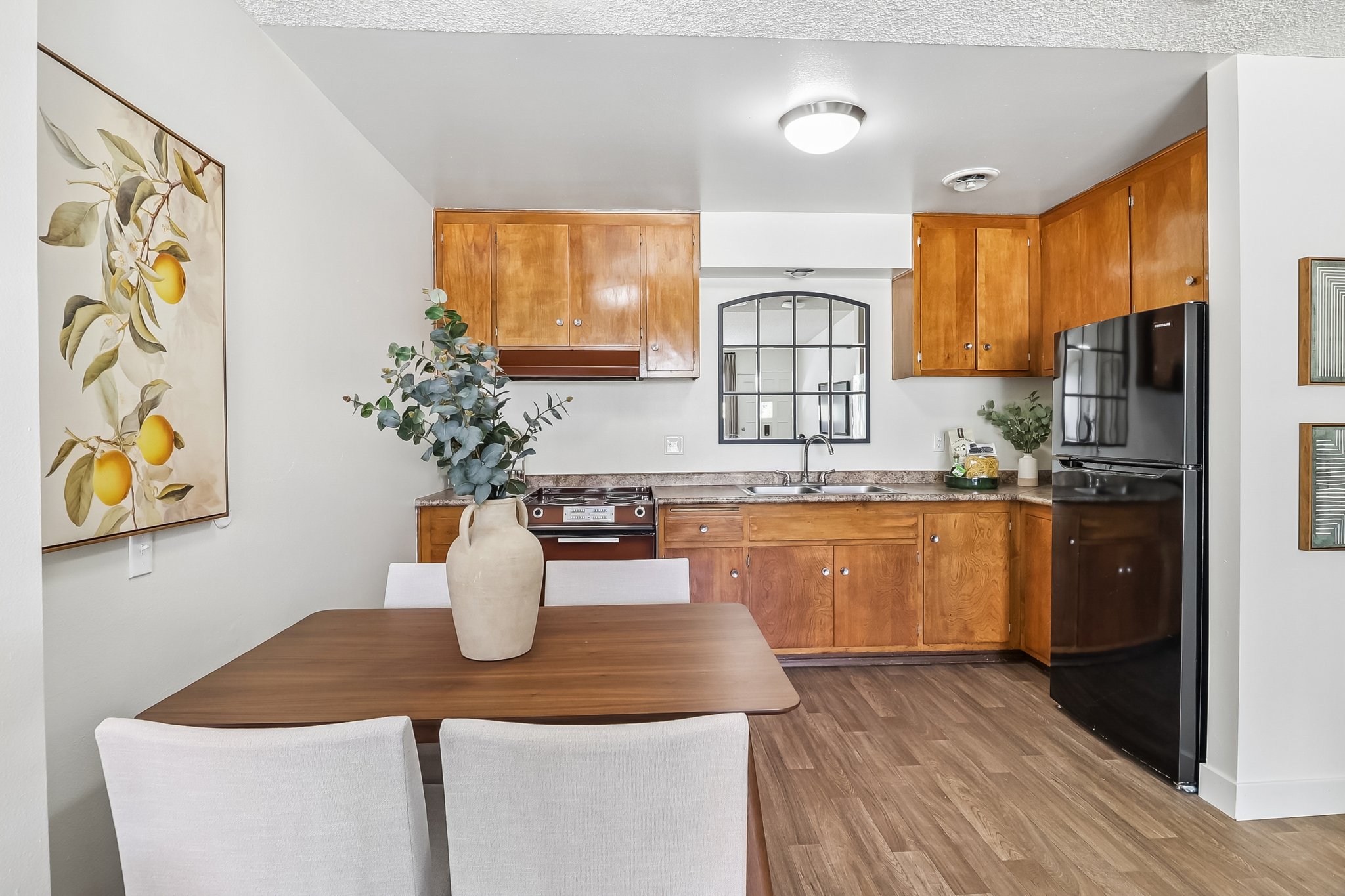 A cozy kitchen with wooden cabinetry, a modern black refrigerator, and a stove. A dining table with white chairs is set in the foreground, and a decorative vase with foliage adds a touch of elegance. A framed artwork featuring lemons hangs on the wall, enhancing the warm ambiance of the space.