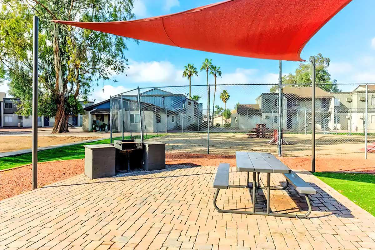 A shaded picnic area featuring a table and a grill, surrounded by a chain-link fence. In the background, there are residential buildings and palm trees, with an open field nearby, providing a spacious outdoor setting.