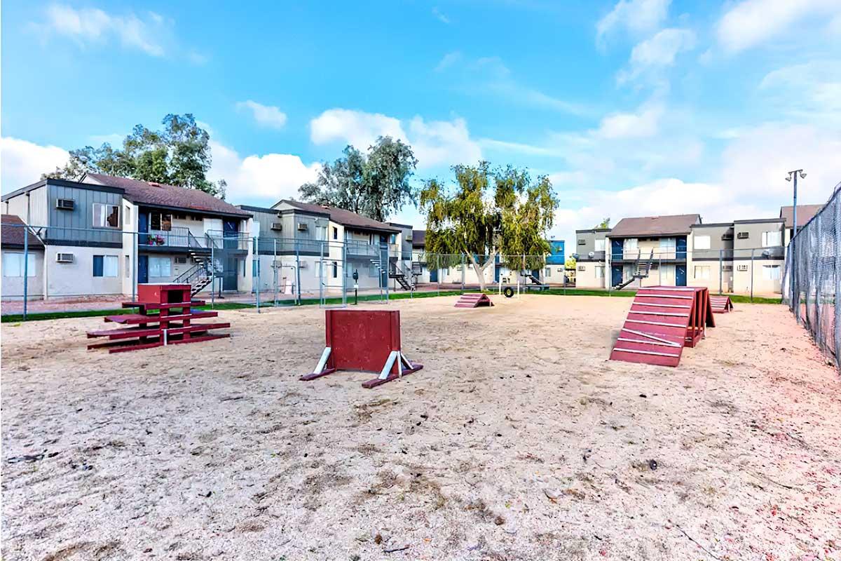 A sandy playground area featuring red agility equipment, including ramps and boxes, surrounded by grassy sections and residential buildings in the background. The sky is partly cloudy, creating a bright and open atmosphere.