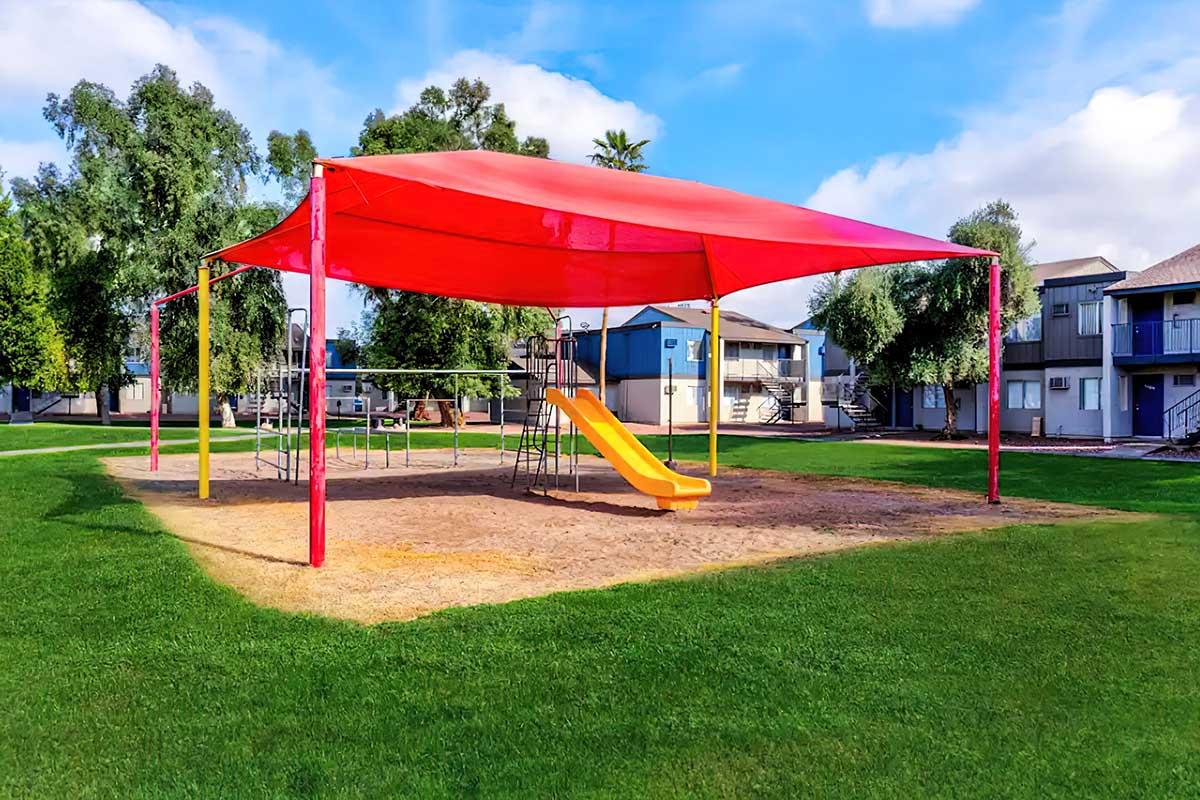 A playground area with a bright red canopy, featuring a yellow slide and swings, situated on a grassy field. In the background, there are residential buildings and trees under a clear blue sky. The scene conveys a cheerful, inviting outdoor space for children to play.