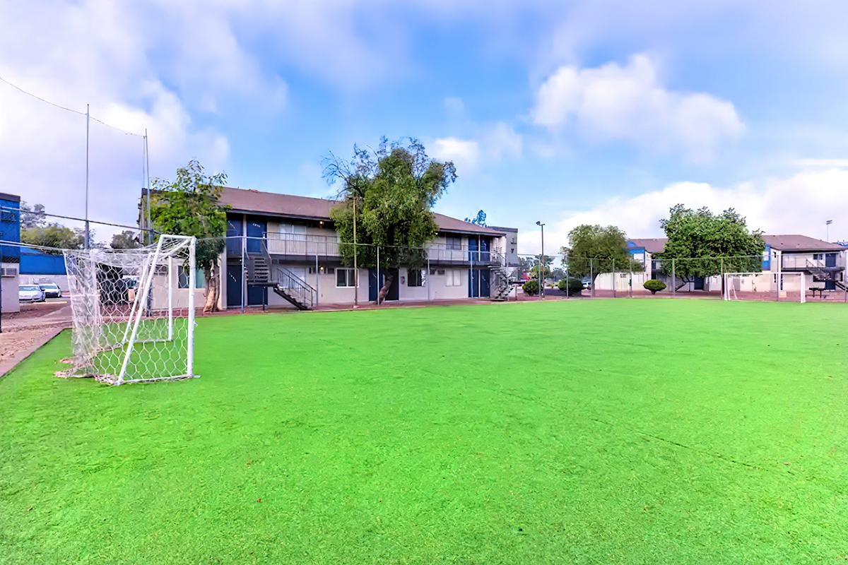 A green soccer field with a goalpost in the foreground, surrounded by residential buildings and green trees under a partly cloudy sky. The field appears well-maintained, providing a recreational space for residents.