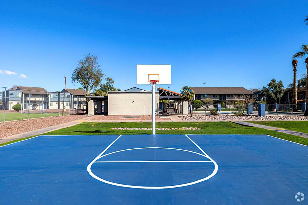 Outdoor basketball court with a blue playing surface, featuring a hoop and backboard. In the background, there are residential buildings and some palm trees, all under a clear blue sky. The area is well-maintained and surrounded by landscaping.