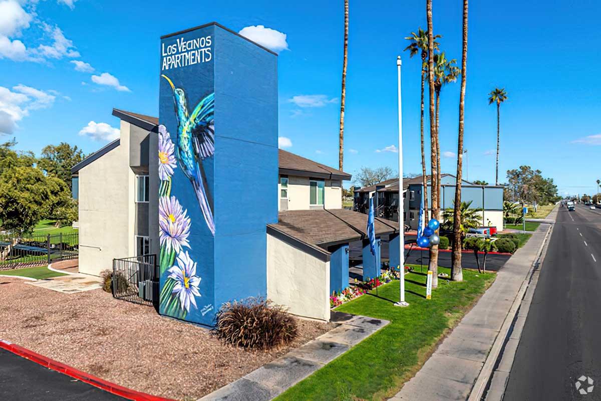 A vibrant mural of a hummingbird and flowers decorates the side of the Los Venados Apartments building. The scene includes palm trees and a clear blue sky, indicating a sunny day. In front, there are flags and balloon decorations, enhancing the welcoming atmosphere of the apartment complex.
