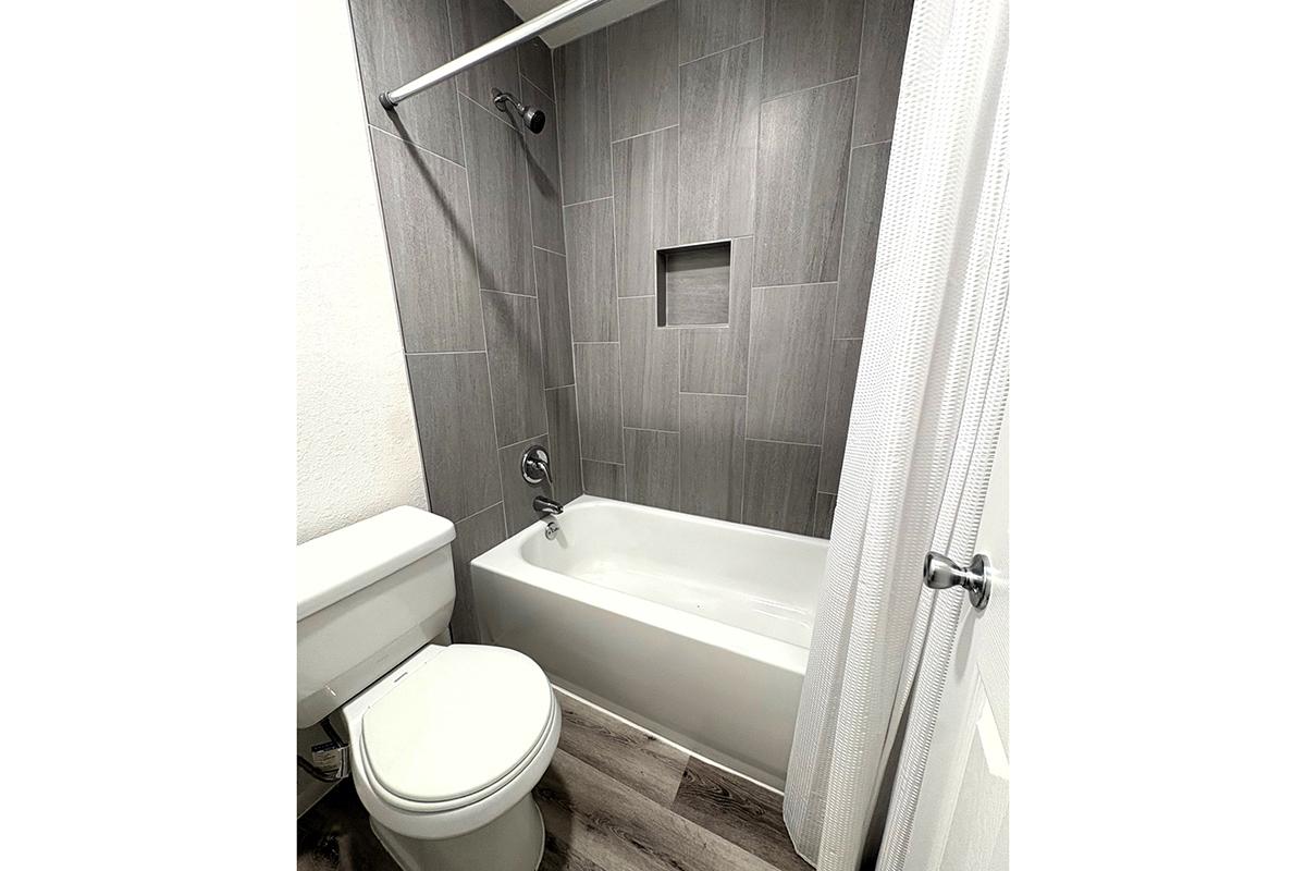A modern bathroom featuring a white bathtub with a showerhead above, a gray tiled wall with a recessed shelf, and a white toilet. The flooring is wood-like, and there is a shower curtain partially visible, adding a clean and contemporary look to the space.