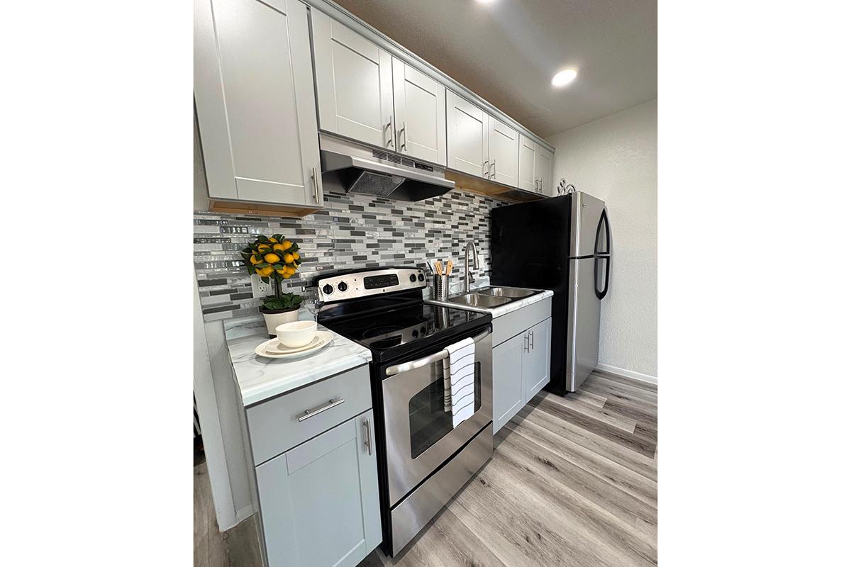 Modern kitchen featuring light gray cabinets, stainless steel appliances, and a stylish backsplash with gray and white tiles. The setup includes a black refrigerator, an oven, and a sink, along with a small vase of yellow flowers on the countertop. Bright lighting enhances the space.
