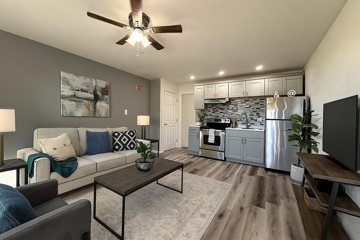 Modern living room and kitchen area in a stylish apartment. Features a light gray sofa with blue accent pillows and a coffee table, alongside a kitchenette with gray cabinets, a stainless steel refrigerator, and a range. Decorative artwork is on the wall, and a ceiling fan is visible above.