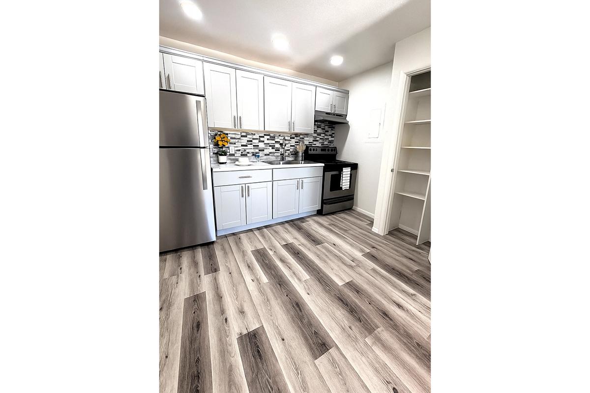 Modern kitchen featuring white cabinetry, stainless steel appliances, and a patterned backsplash. The floor is light wood-look laminate, and there is a large pantry on the right. Natural light enhances the spacious feel of the room.