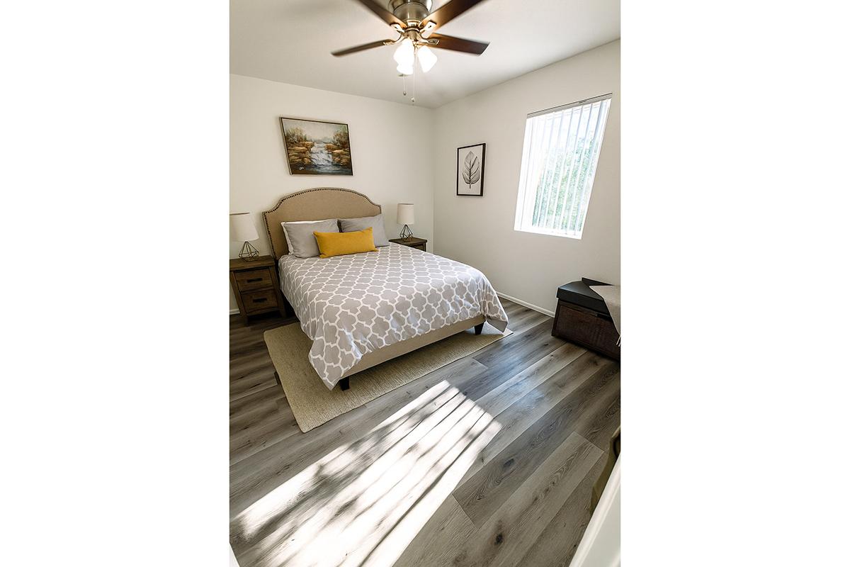 A cozy bedroom featuring a queen-sized bed with a light gray geometric patterned comforter, yellow accent pillow, and wooden nightstands on either side. Natural light enters through a vertical blind window, illuminating the room's neutral decor and wooden floor. A framed landscape painting hangs on the wall.