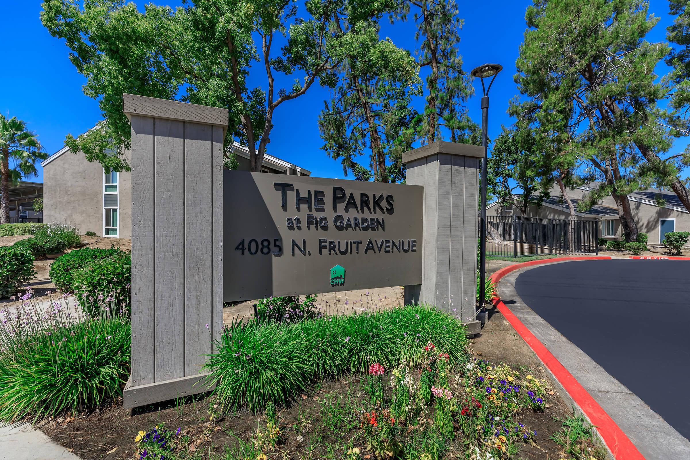 Sign displaying "The Parks at Fig Garden" with the address "4085 N. Fruit Avenue," surrounded by landscaped greenery and colorful flowers, under a clear blue sky.
