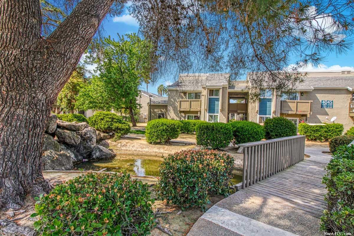 A serene outdoor scene featuring a wooden footbridge over a small pond, surrounded by lush greenery and manicured bushes. In the background, there are multi-story buildings with balconies, framed by trees, creating a peaceful residential atmosphere. The sky is bright with scattered clouds.
