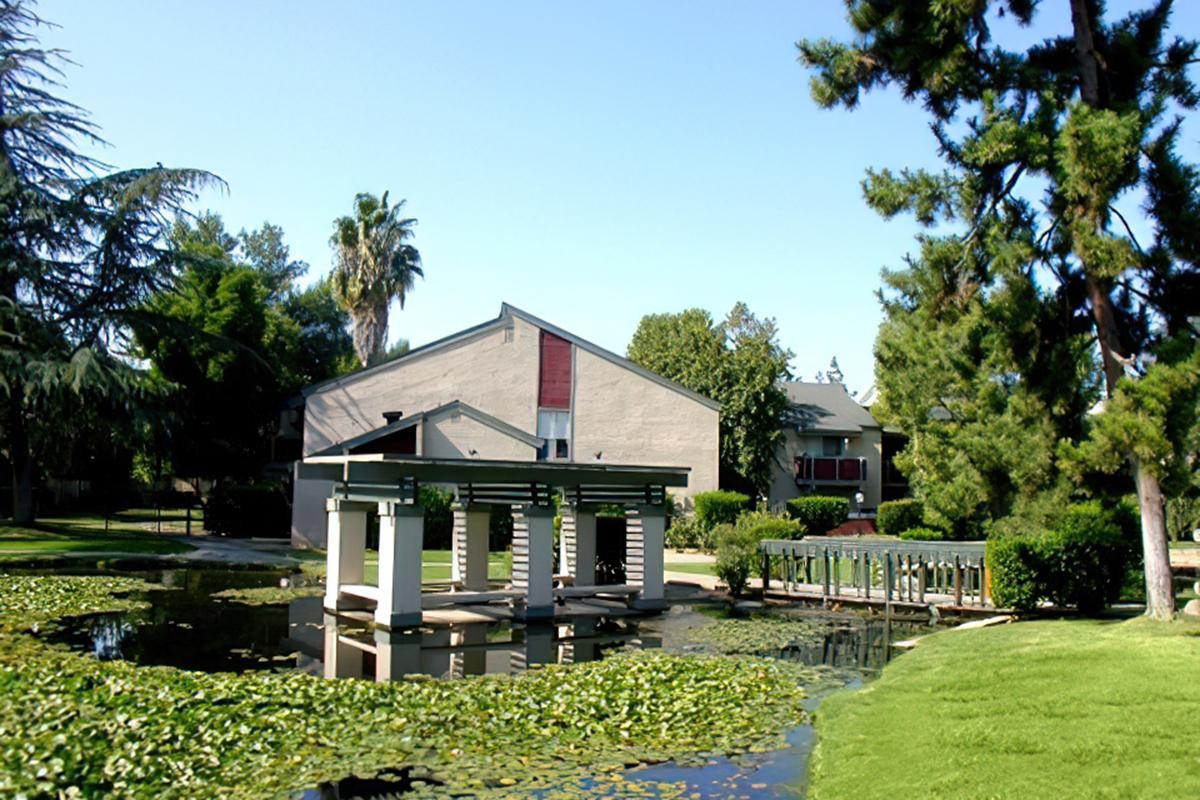  A serene park scene featuring a small pond surrounded by greenery. In the foreground, a covered gazebo sits on the water's edge, reflecting in the pond.