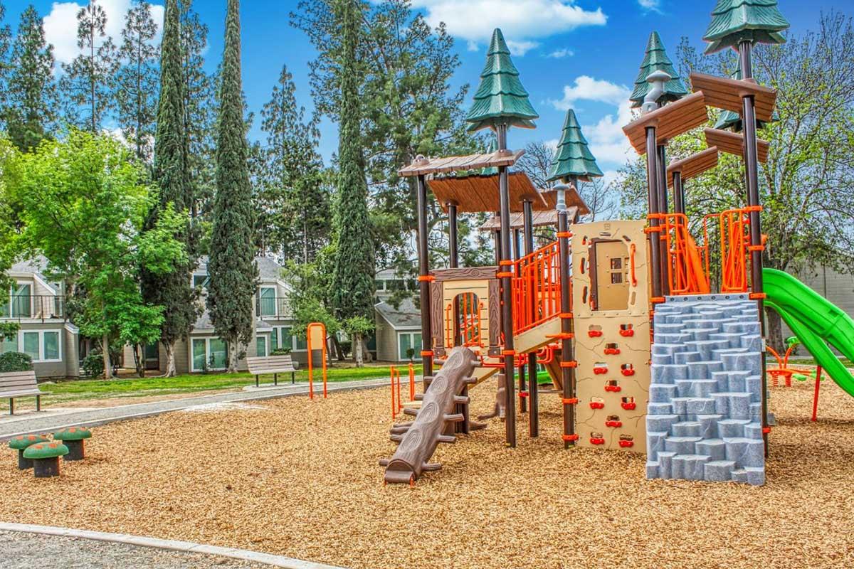 A colorful playground featuring a climbing structure, slides, and climbing walls, surrounded by wood chips and tall trees. In the background, there are residential buildings with green spaces. The sky is blue with scattered clouds.