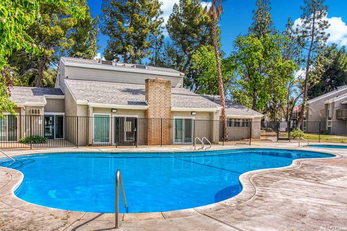 A clear blue swimming pool surrounded by a paved area, with a modern building in the background. The building features large windows and a chimney, set among lush green trees. The scene is bright and inviting, perfect for relaxation and leisure.