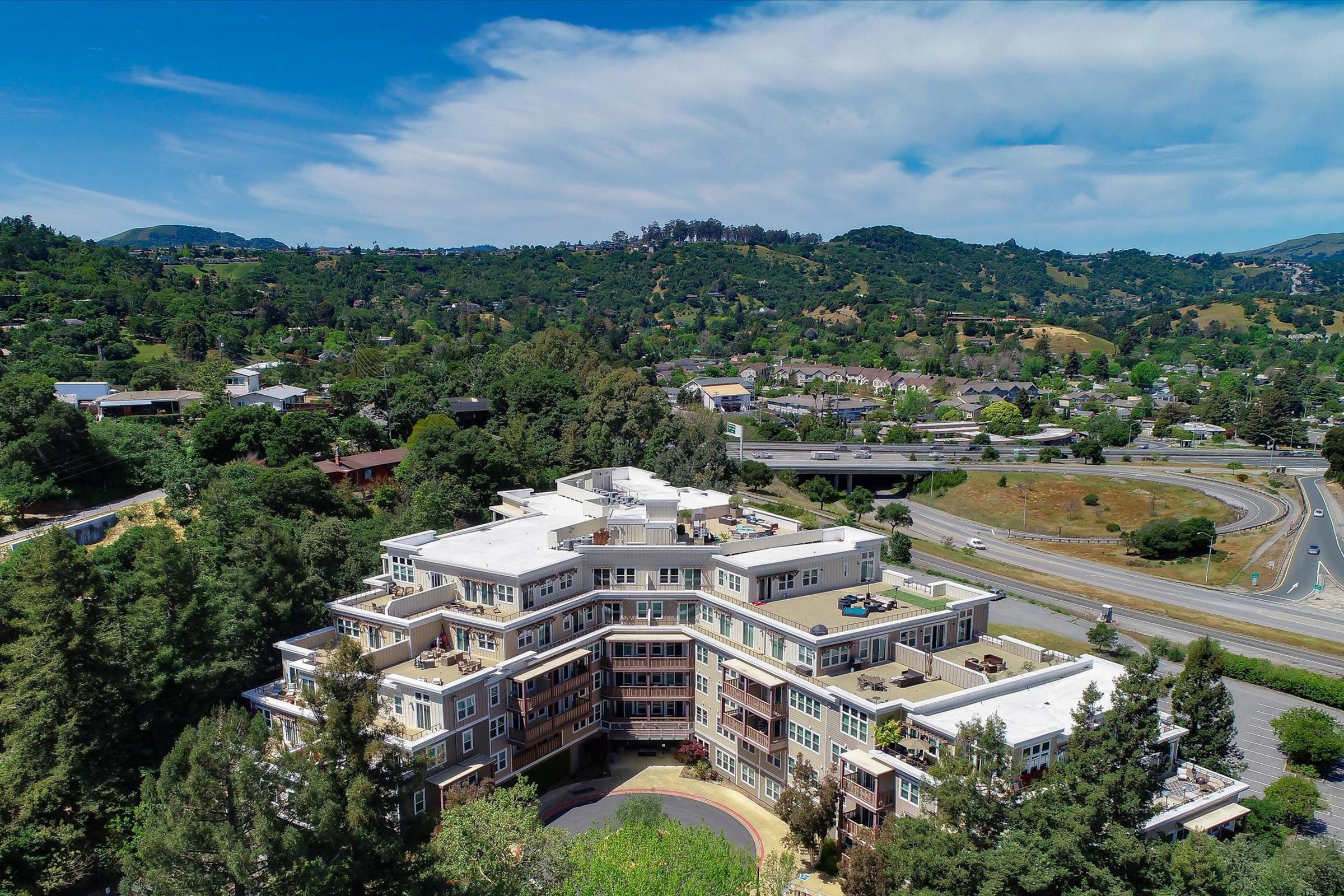 Aerial view of a modern multi-story building surrounded by lush green hills and trees, with a highway running nearby. The sky is clear with a few clouds, highlighting the natural landscape and urban development.