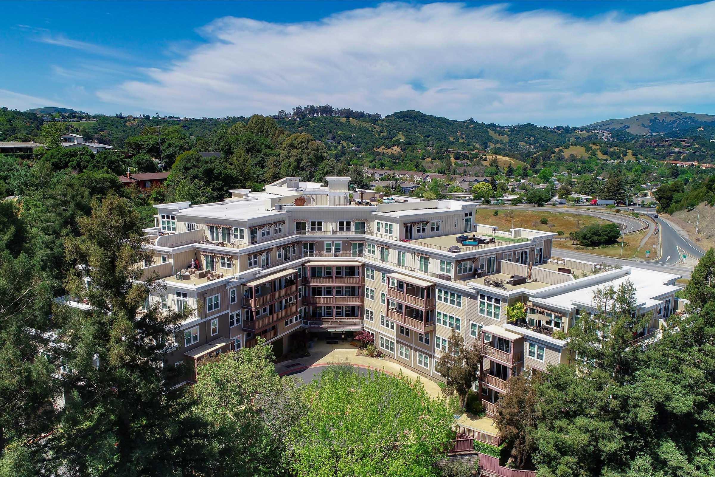 Aerial view of a modern multi-story apartment complex surrounded by greenery and mountains. The building features a contemporary design with balconies and large windows. In the background, hills and a highway can be seen, emphasizing the scenic location.