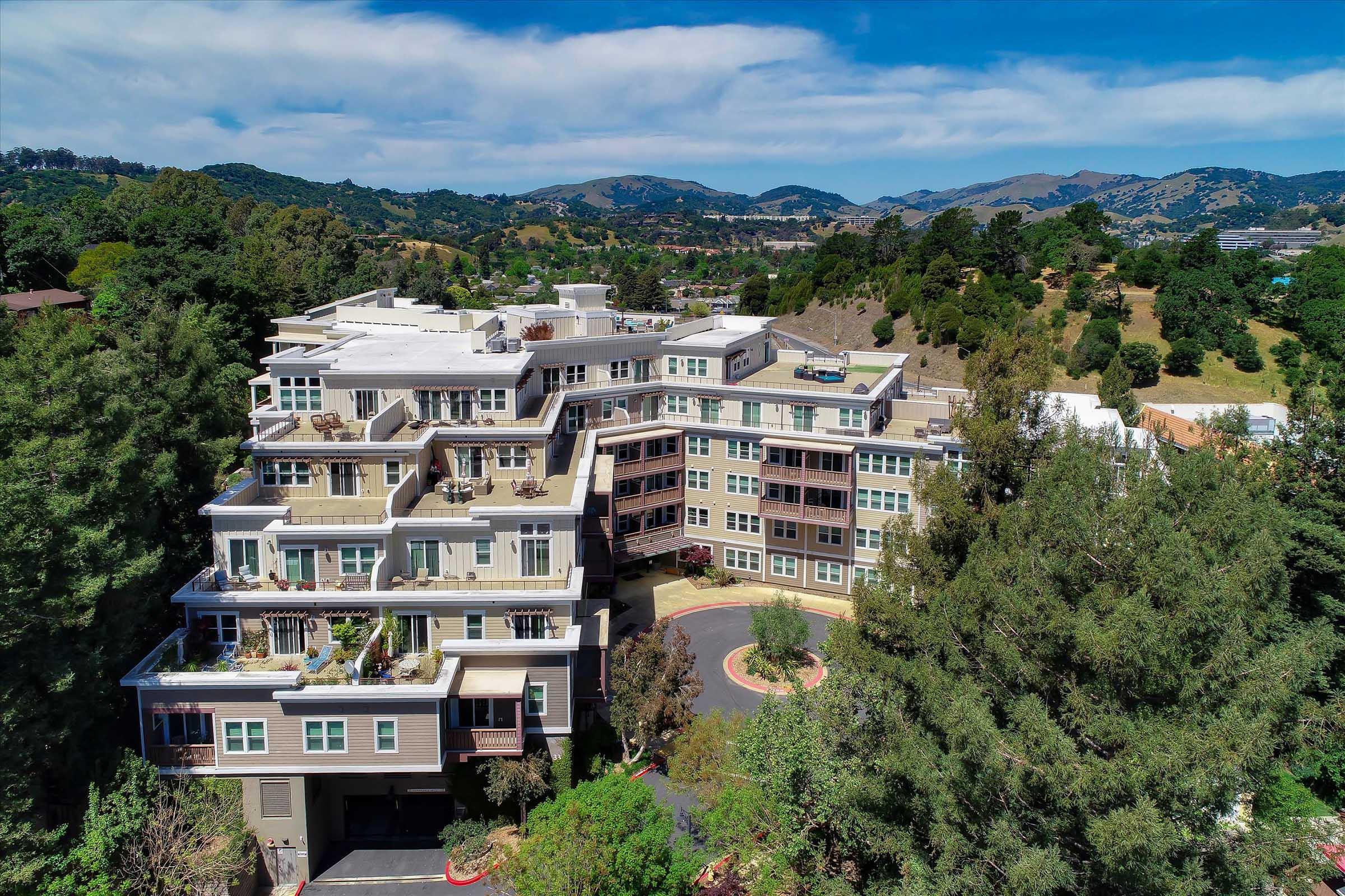 Aerial view of a multi-story residential building surrounded by trees and rolling hills. The building features balconies and a landscaped area, with a winding road leading to its entrance. The sky is partly cloudy, and the mountains are visible in the background.