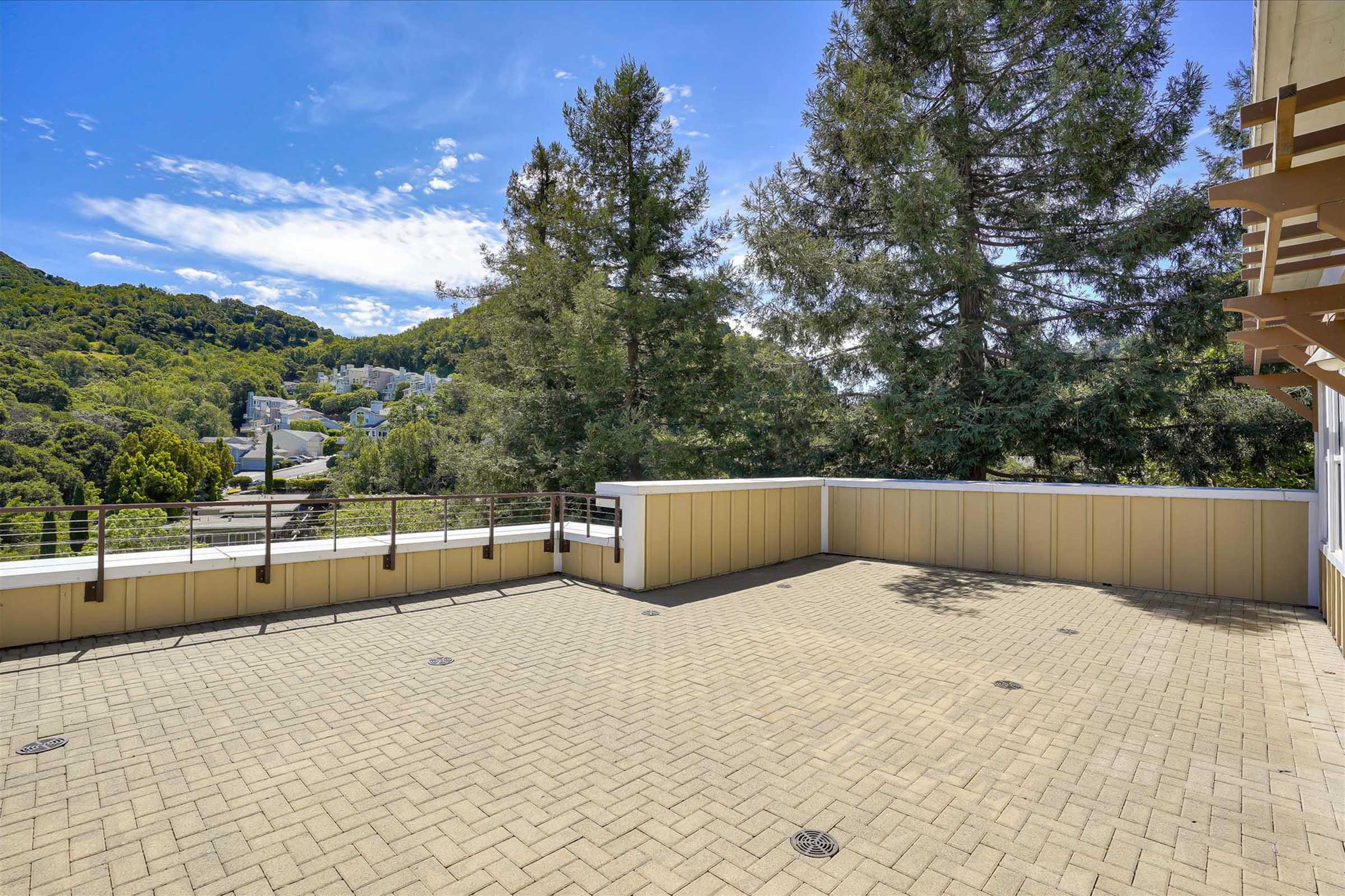 A spacious outdoor terrace featuring a tiled floor, surrounded by green trees and distant hills under a blue sky with scattered clouds. The railing offers a view of the scenic landscape, creating an inviting space for relaxation or gatherings.