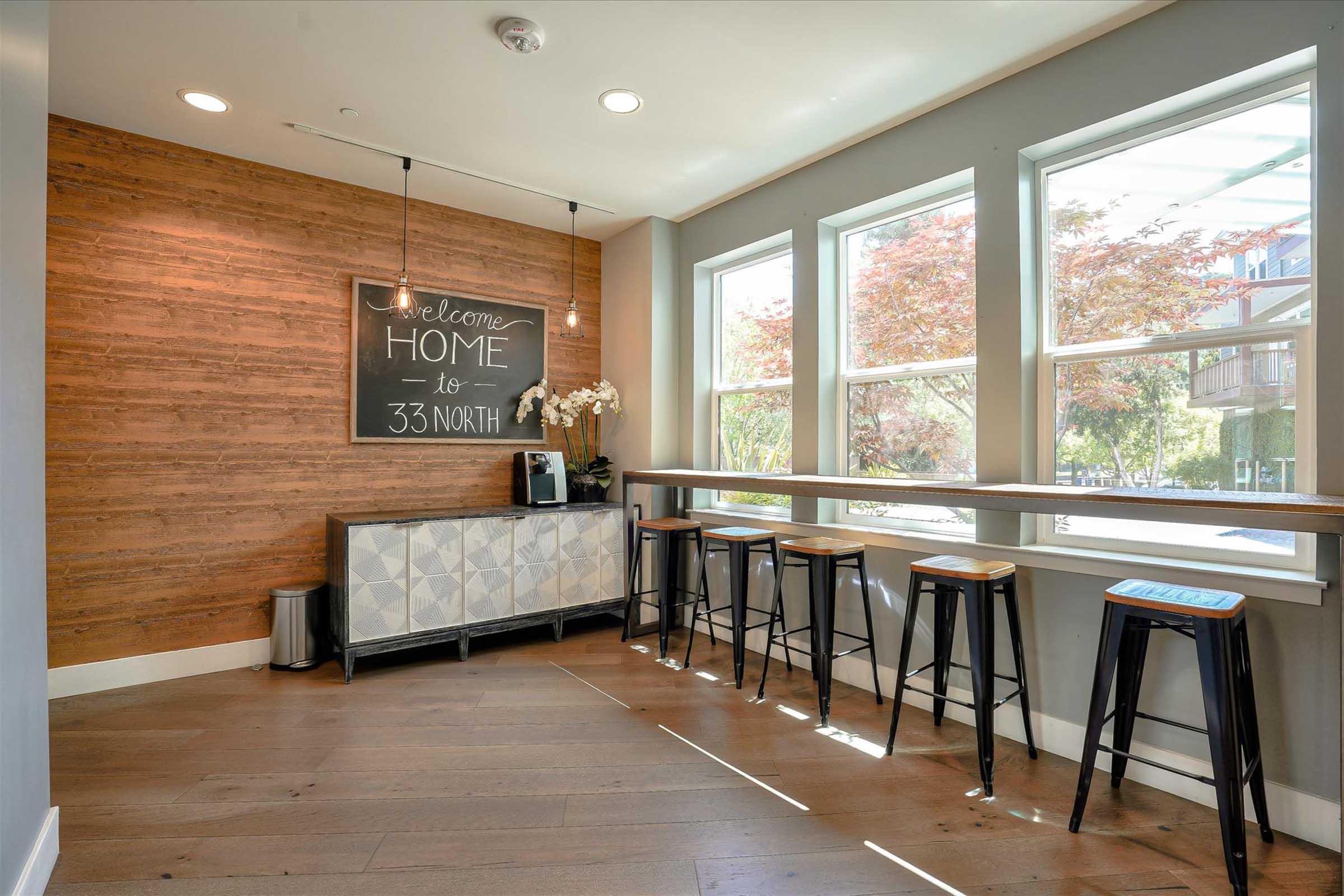A modern interior space featuring a welcoming sign that reads "Welcome Home to 33 North." It includes a wooden accent wall, a gray and white patterned cabinet, and tall black stools arranged at a long, narrow table next to large windows letting in natural light.