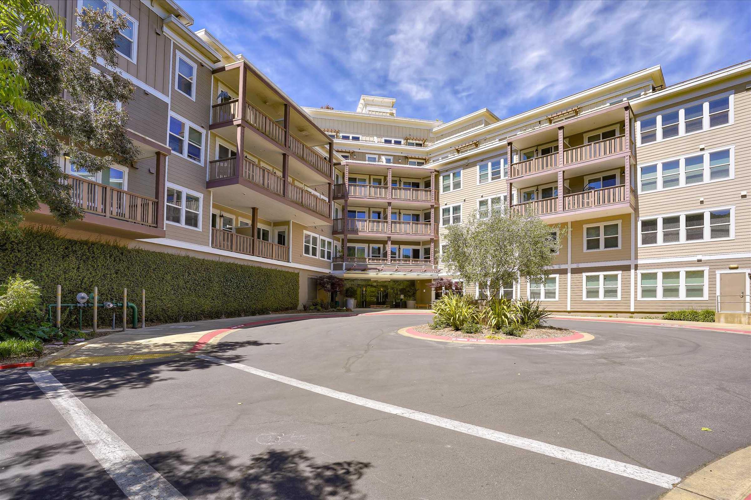 A modern multi-story apartment building with a circular driveway, surrounded by greenery. The architecture features large windows and balconies. The sky is partly cloudy, creating a bright and inviting atmosphere.