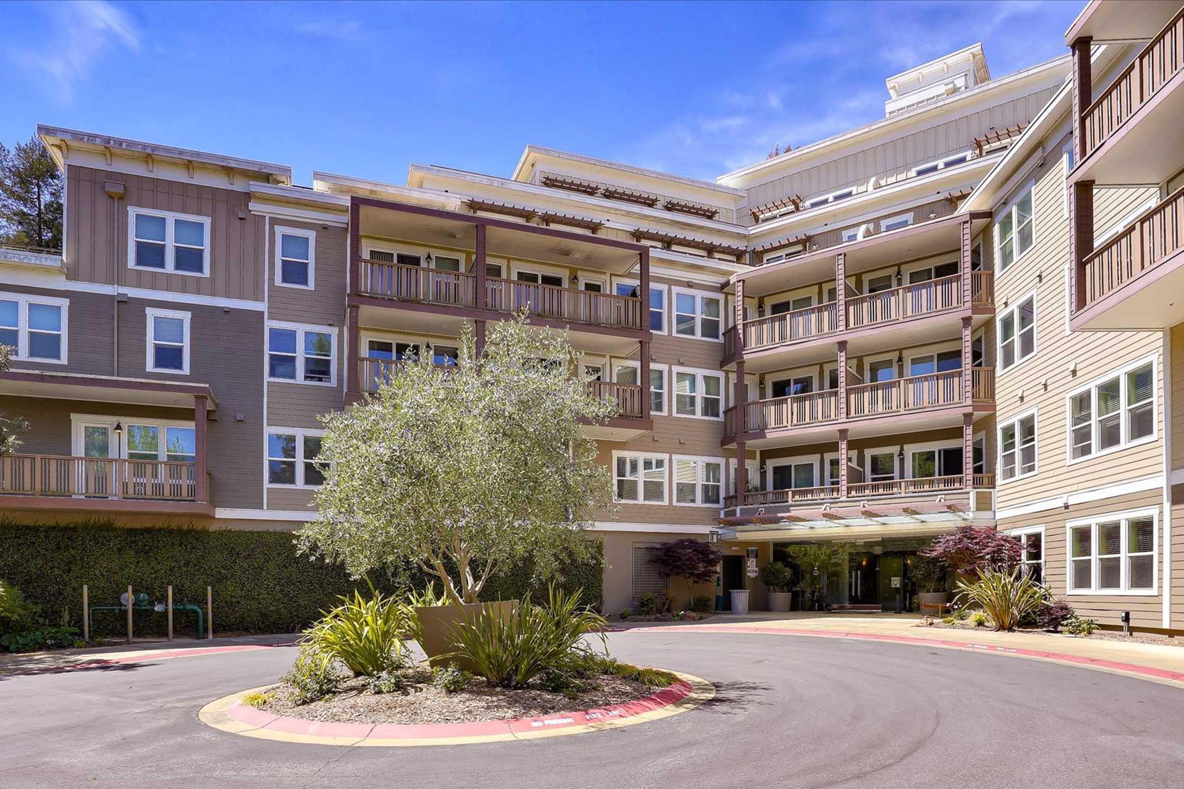 Modern apartment building with multiple balconies and large windows, surrounded by greenery and landscaping. The entrance features a driveway curving around a central garden area with plants, creating an inviting atmosphere. The structure has a mix of light and dark-colored siding under a clear blue sky.