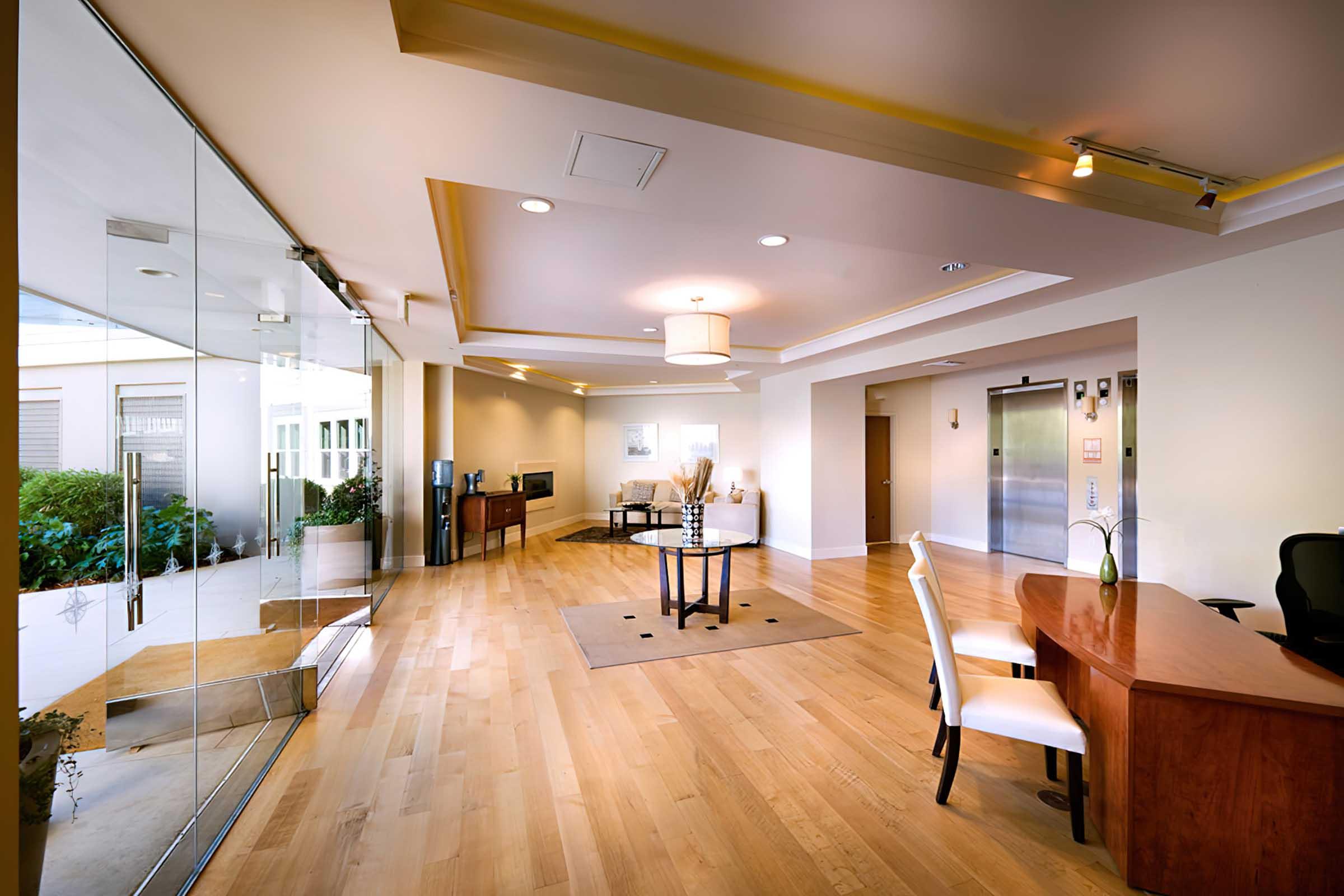 Modern interior view of a spacious lobby featuring wooden flooring, a glass wall, a small seating area with chairs, a round table, and a reception desk. Soft lighting illuminates the area, and plants are visible outside through the glass. The design is contemporary and inviting.