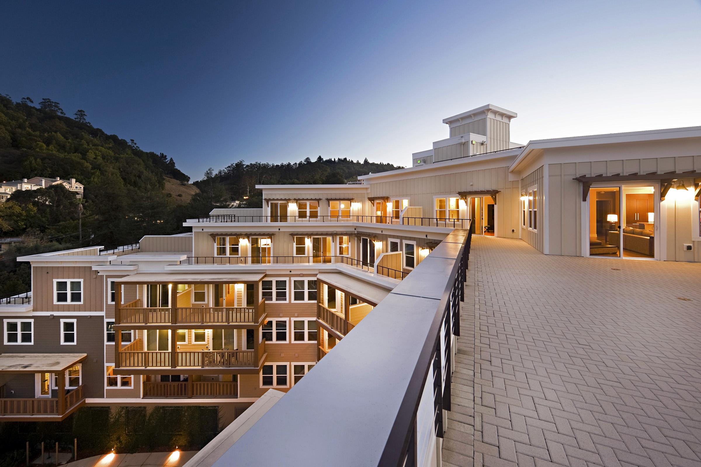 A modern multi-story building with a rooftop terrace, featuring an expansive balcony overlooking a landscaped area. The architecture includes a mix of light-colored and dark wooden facades, with multiple windows illuminated at dusk, set against a backdrop of rolling hills and trees.