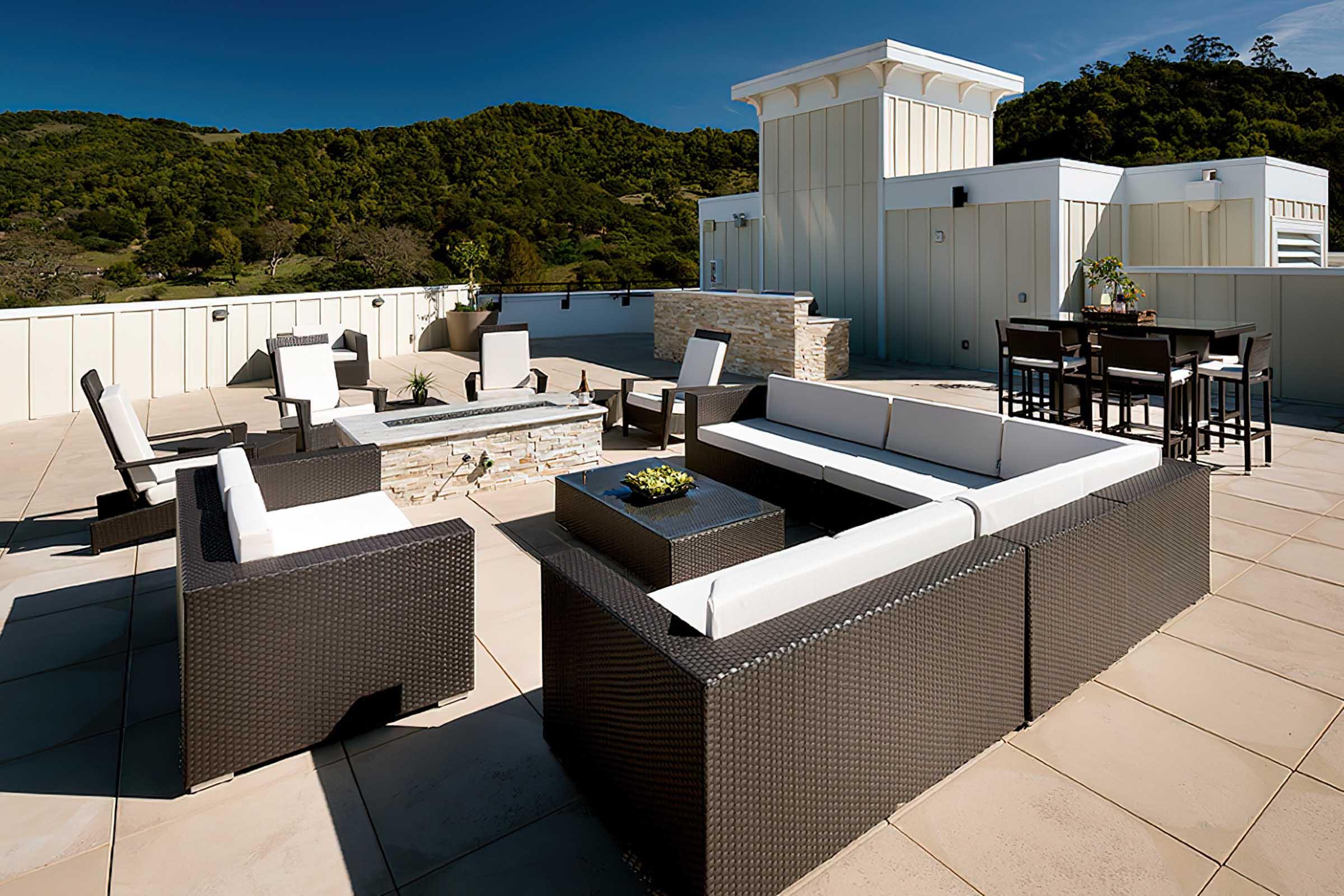 A modern rooftop patio featuring sleek outdoor furniture, including white cushioned sofas, a stone firepit, and a dining table. The space is surrounded by greenery and has a clear blue sky overhead, creating a stylish and inviting outdoor living area.