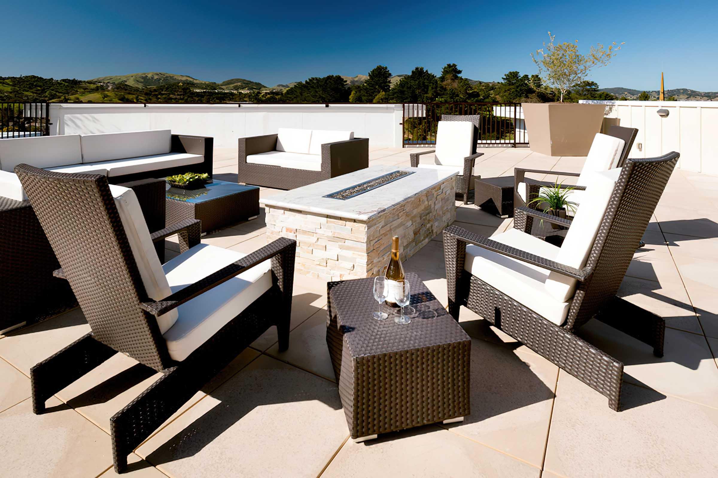A modern outdoor seating area featuring elegant black wicker furniture with white cushions. The setup includes a central stone fire pit table, surrounded by armchairs and small side tables. The backdrop showcases a clear blue sky and rolling green hills, creating a serene atmosphere for relaxation.