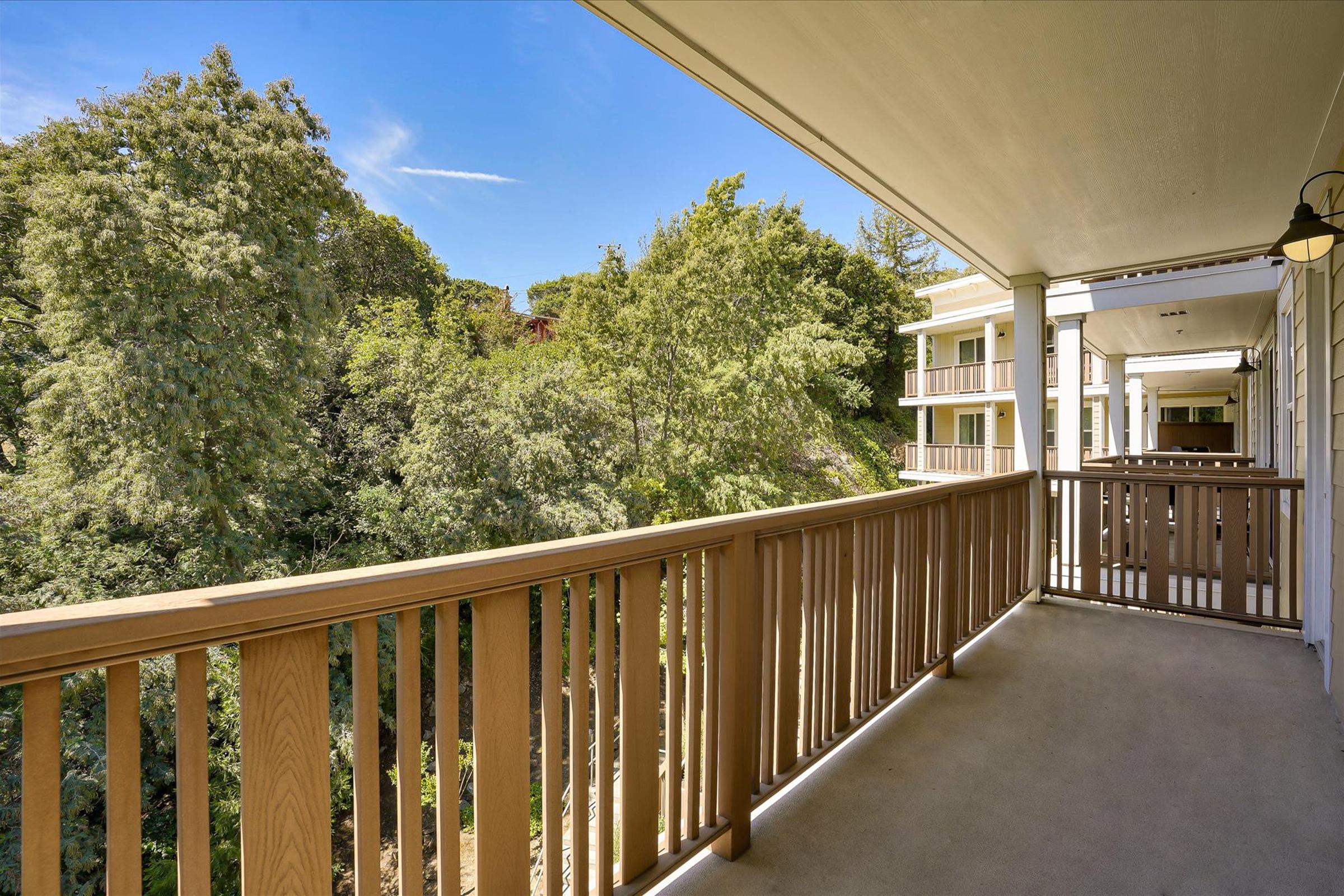 A view from a covered walkway or balcony with wooden railings, showing green trees and landscaping in the background. The sky is clear with a few clouds, and the scene is bright and inviting, suggesting a peaceful outdoor setting near a building.