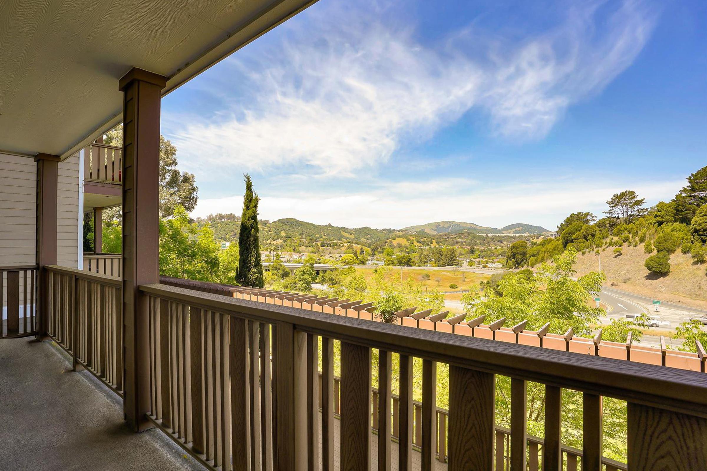 View from a balcony overlooking a scenic landscape. Lush green hills and trees are visible under a blue sky with wispy clouds. The foreground features a wooden railing, and the overall atmosphere conveys a peaceful, natural setting.